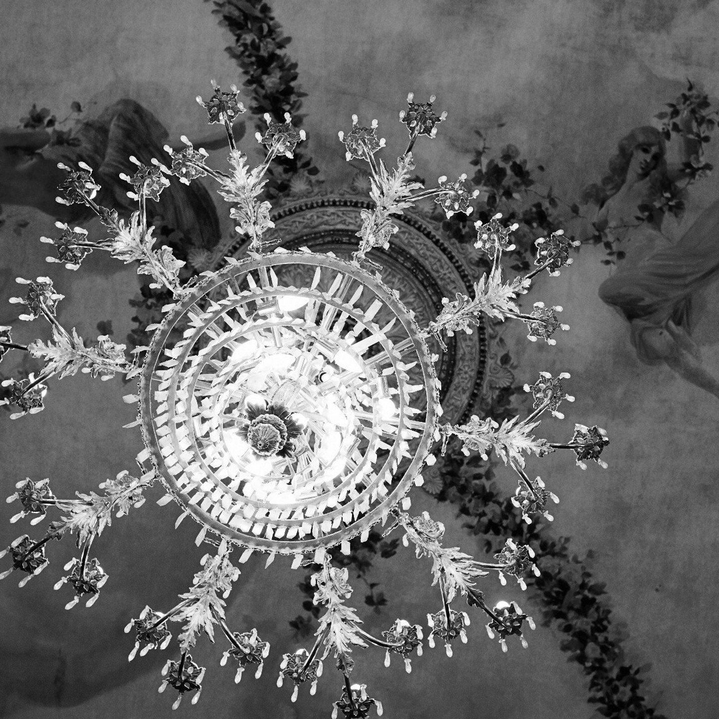 Black and white image of crystal chandelier with ceiling paintings in the background.