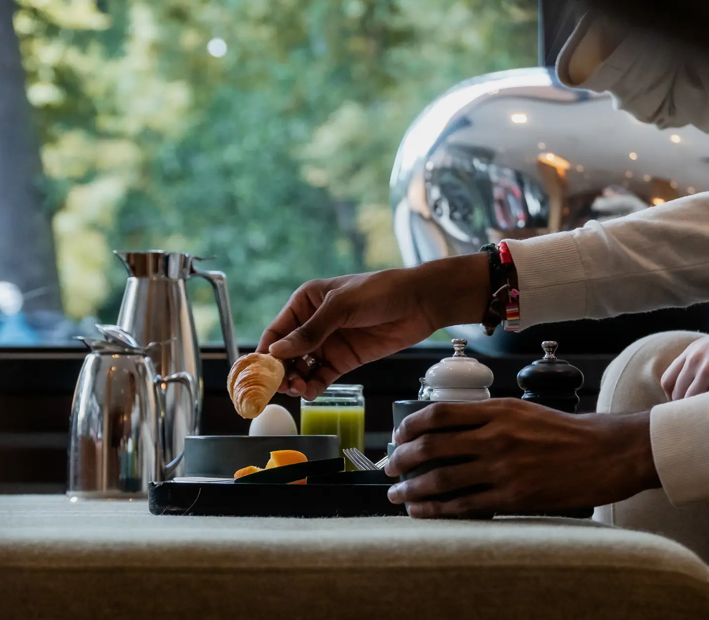 Person serverar frukost med kaffe, croissant och kokt ägg vid ett bord med naturligt ljus utifrån ett fönster