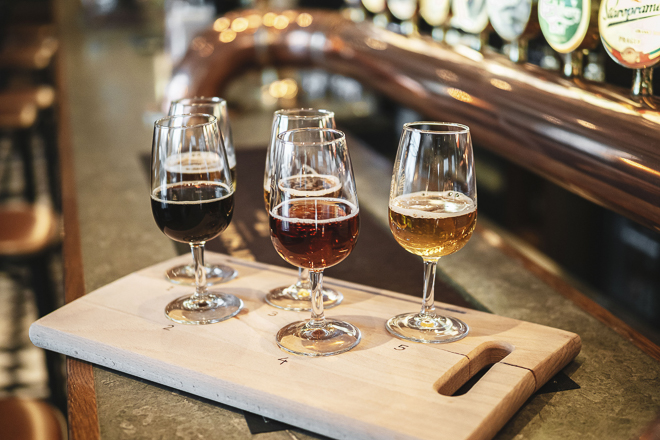 Fyra smakprovsglas med öl på träbricka, placerade på en bar med koppardetaljer och ölkranar i bakgrunden.