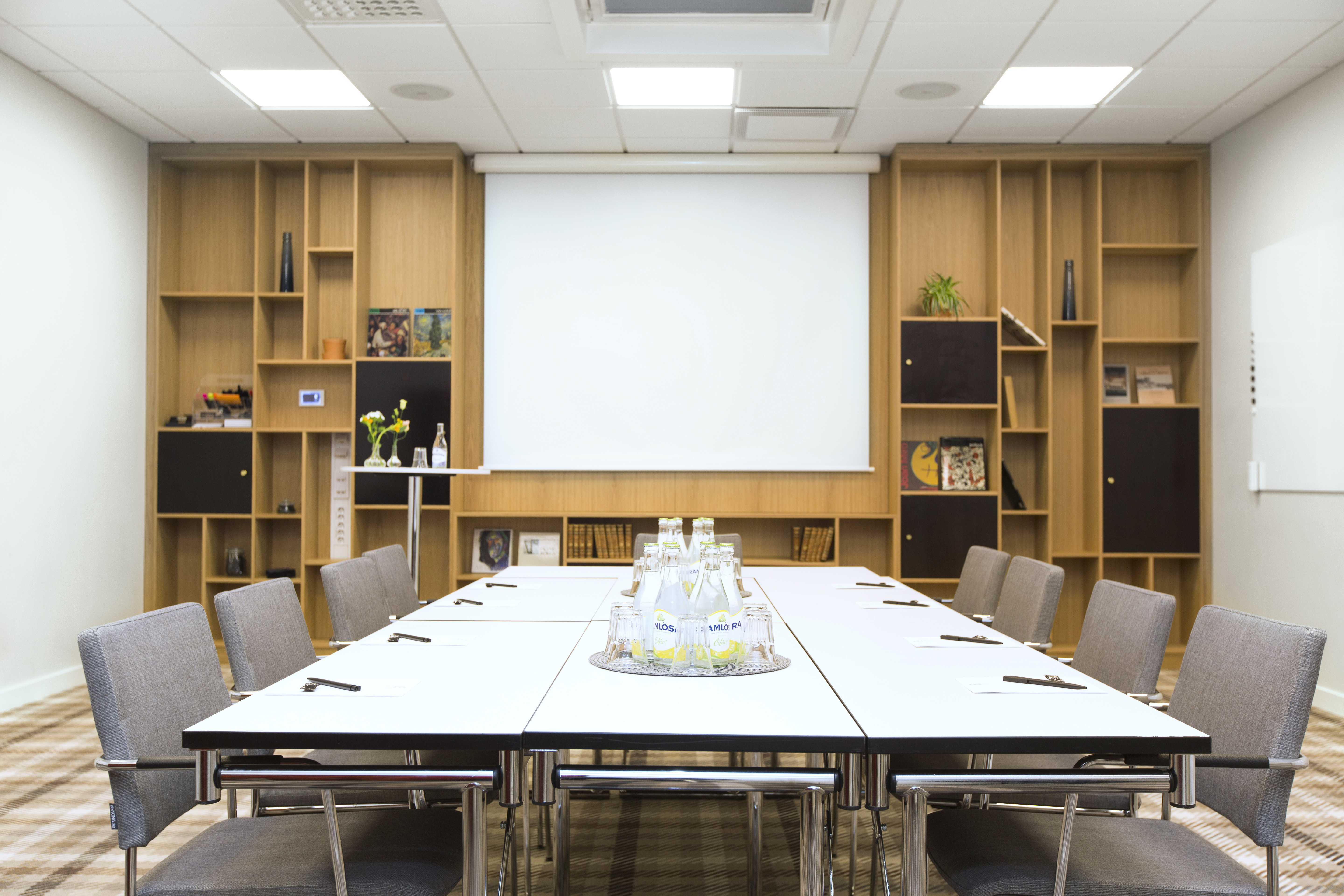 Konferensrum med ett stort bord omgivet av grå stolar, ett projiceringsduk och moderna hyllor i trä fyllda med böcker och dekorationer i en ljus och välorganiserad miljö.