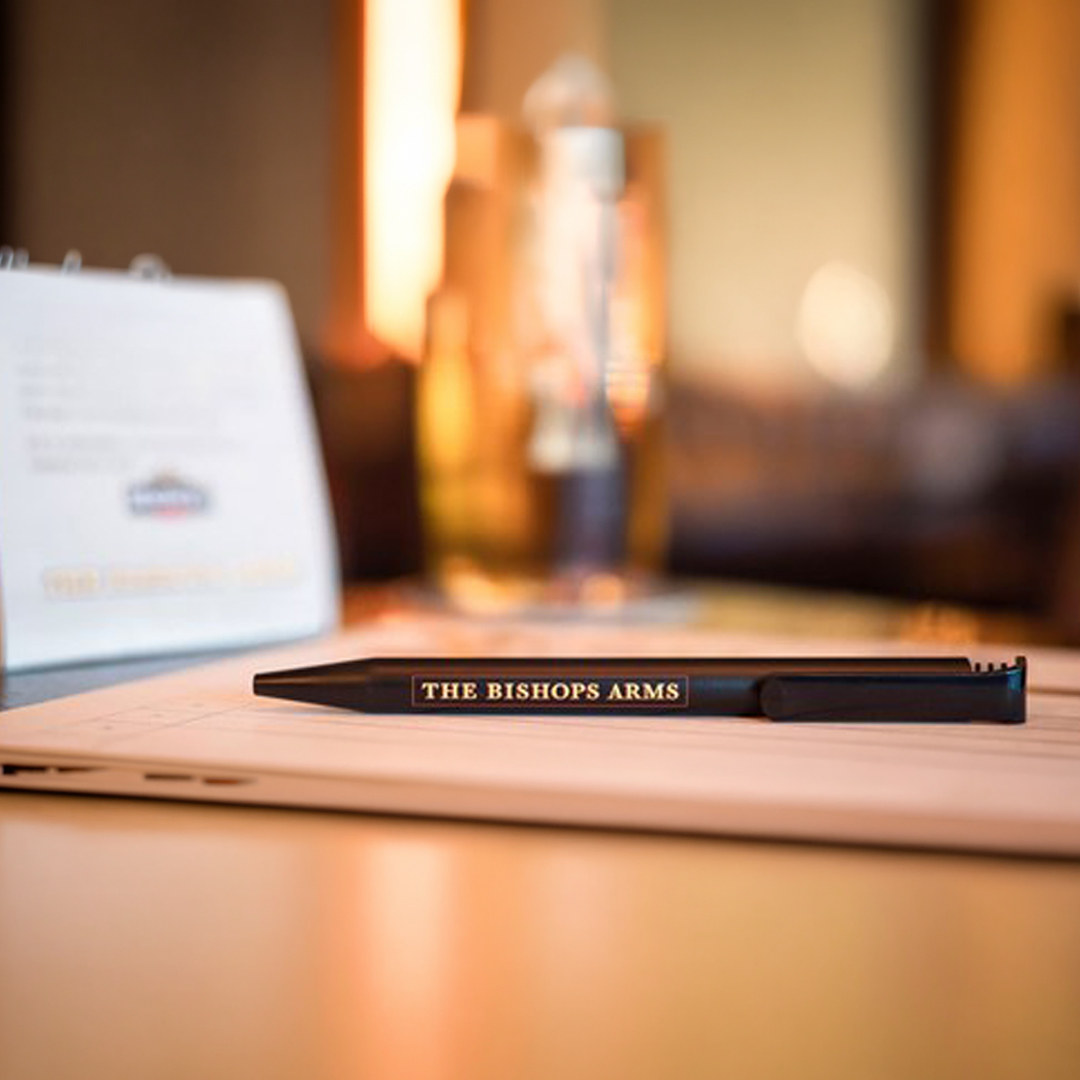 Svart penna med texten "The Bishops Arms" placerad på ett bord med ett glas och ett menyhäfte i bakgrunden, i en varm och inbjudande miljö.