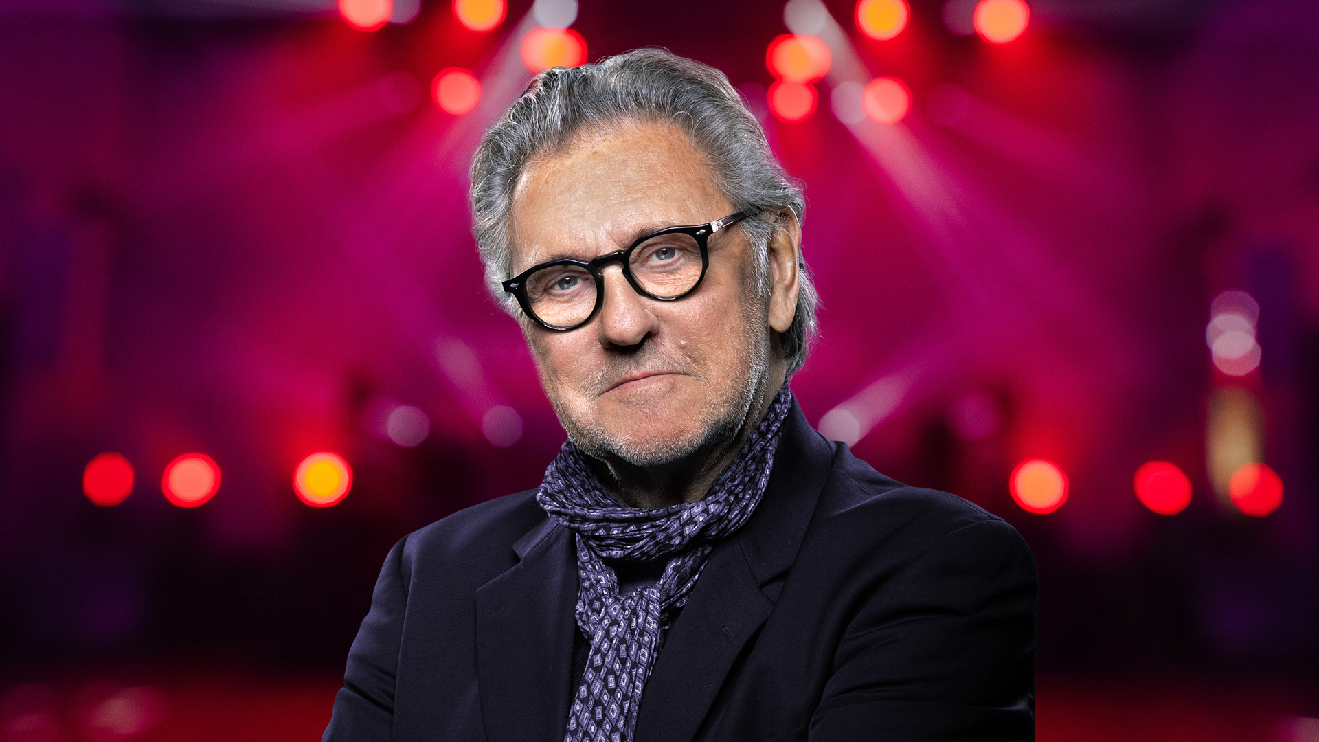 Man med glasögon och scarf i en mörk kostym med suddig, färgstark scenbakgrund.