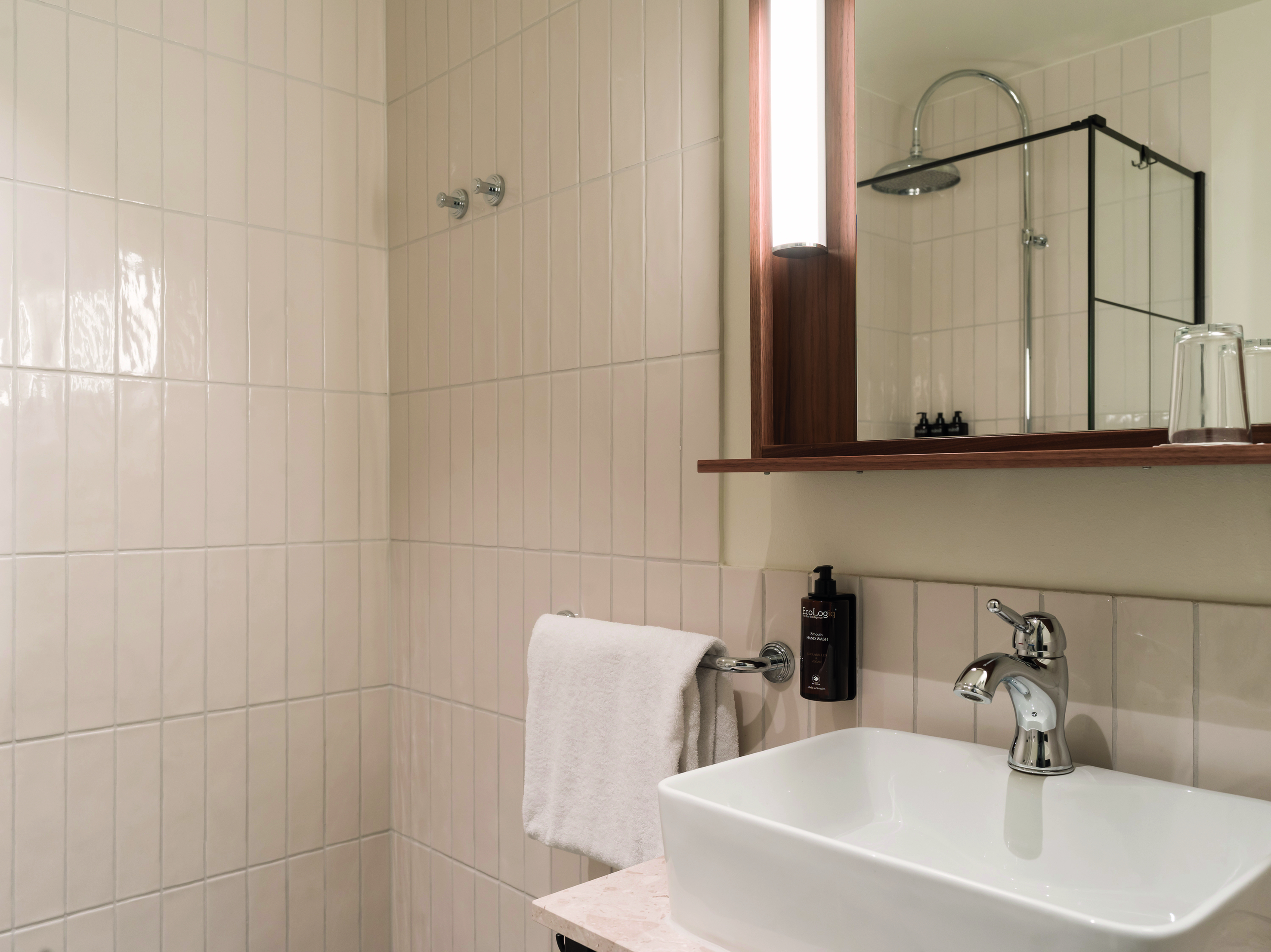 Modernt badrum med vita kakelväggar, rektangulär handfat, och spegel med inbyggd belysning. Silverfärgad kran och dusch, samt handdukshängare med vit handduk