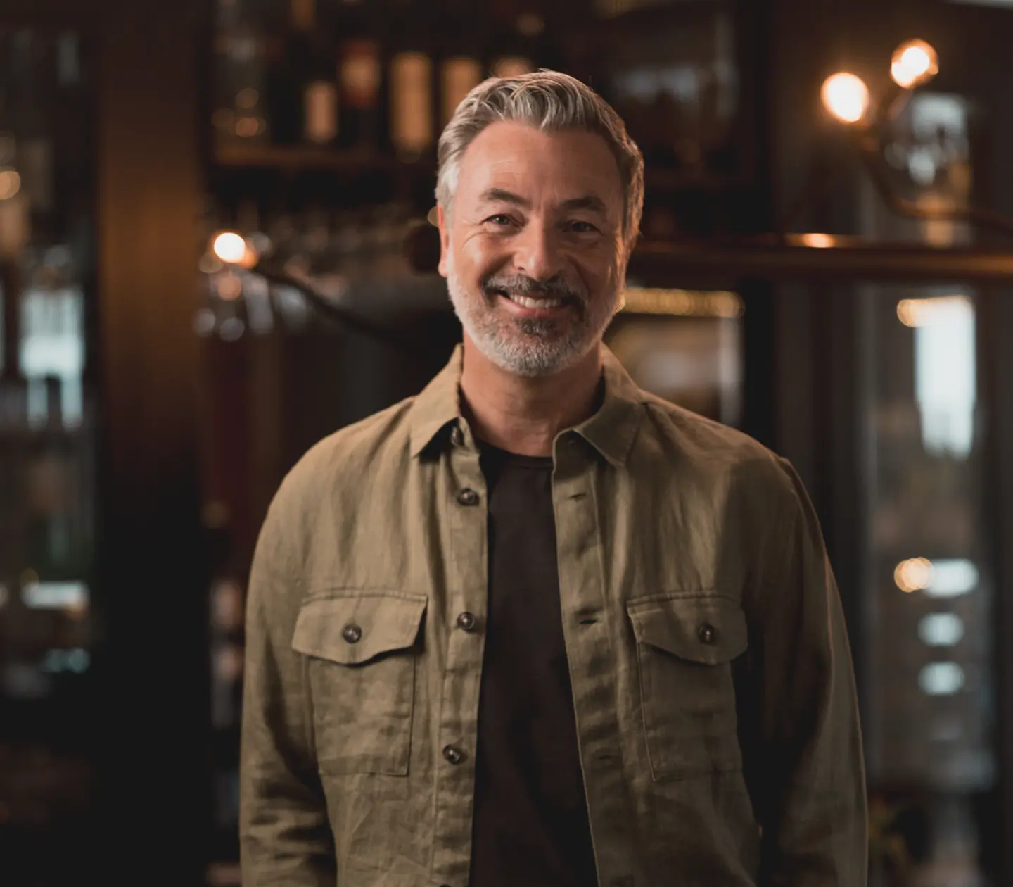 Smiling man wearing a green jacket in a dimly lit modern bar.