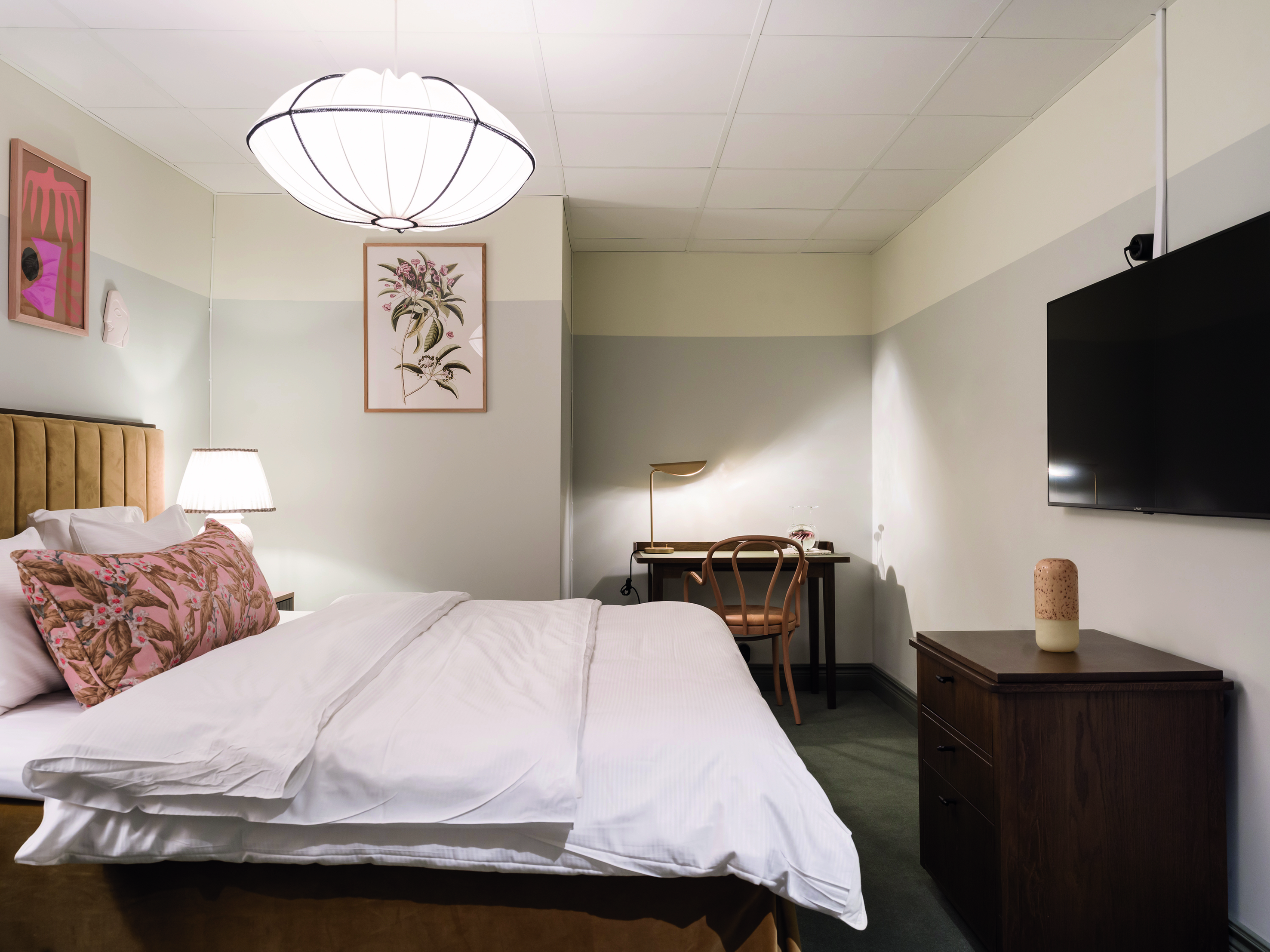 Modernt hotellrum med dubbelsäng, skrivbord och väggmonterad TV. Harmonisk inredning med pastellfärger och dekorativa kuddar. Optimal belysning med stilfull taklampa och bordslampa för en avslappnad atmosfär