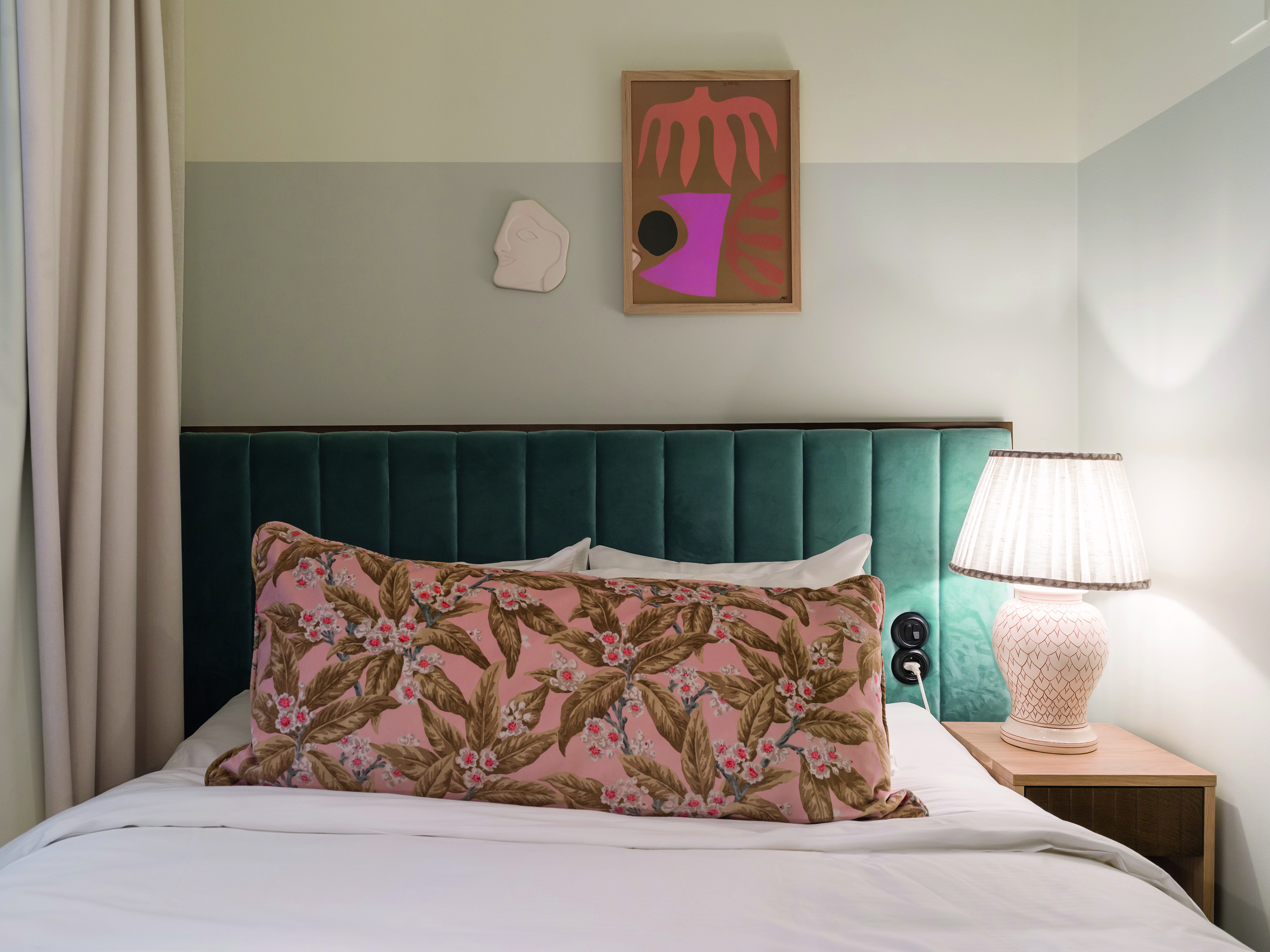 Sovrum med en elegant säng med grön huvudgavel och blommig kudde, samt en sidobordslampa och konst på väggen