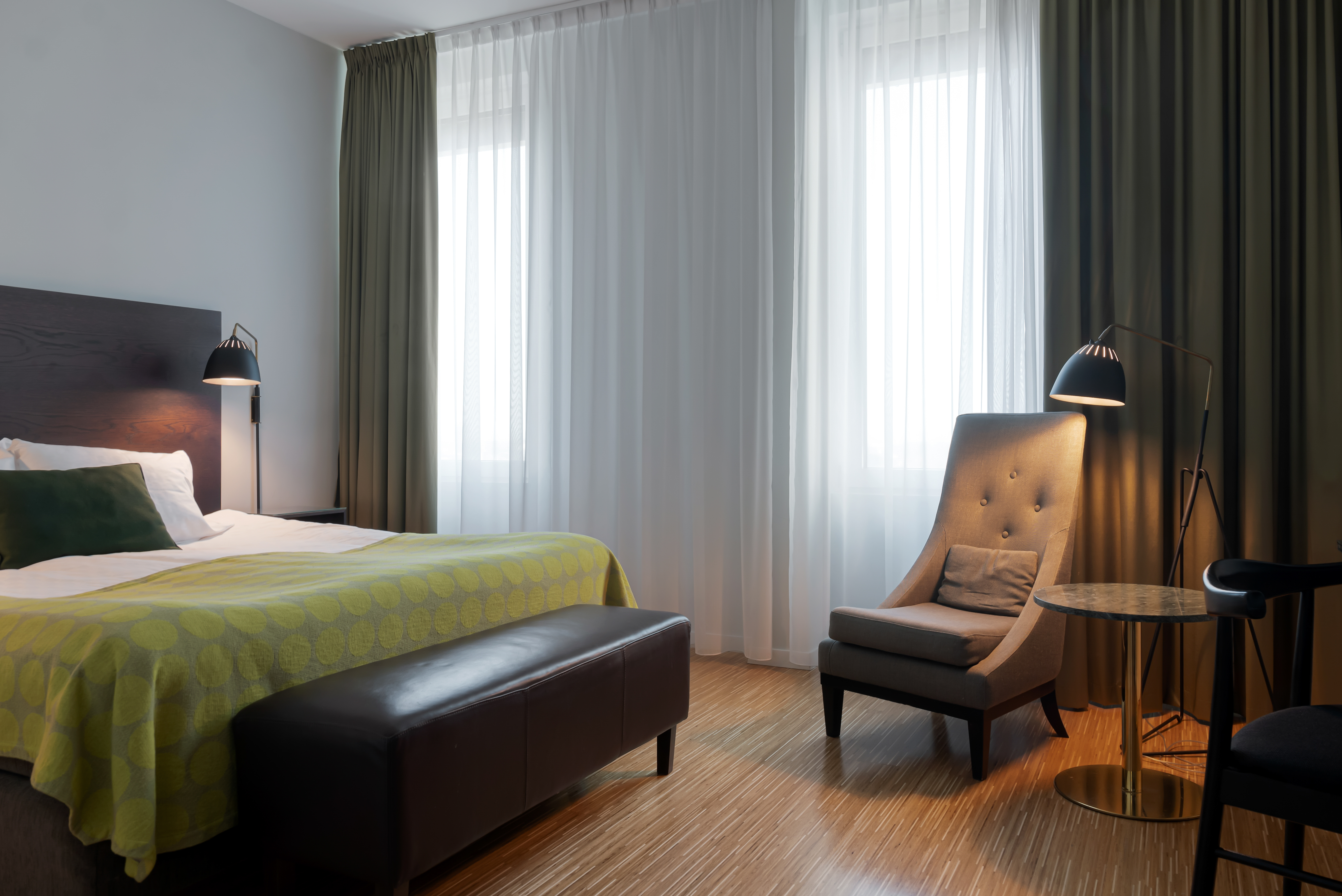 Modern hotel room with a comfortable bed, stylish chair, and soft lighting, featuring a neutral color palette and large windows with sheer curtains.