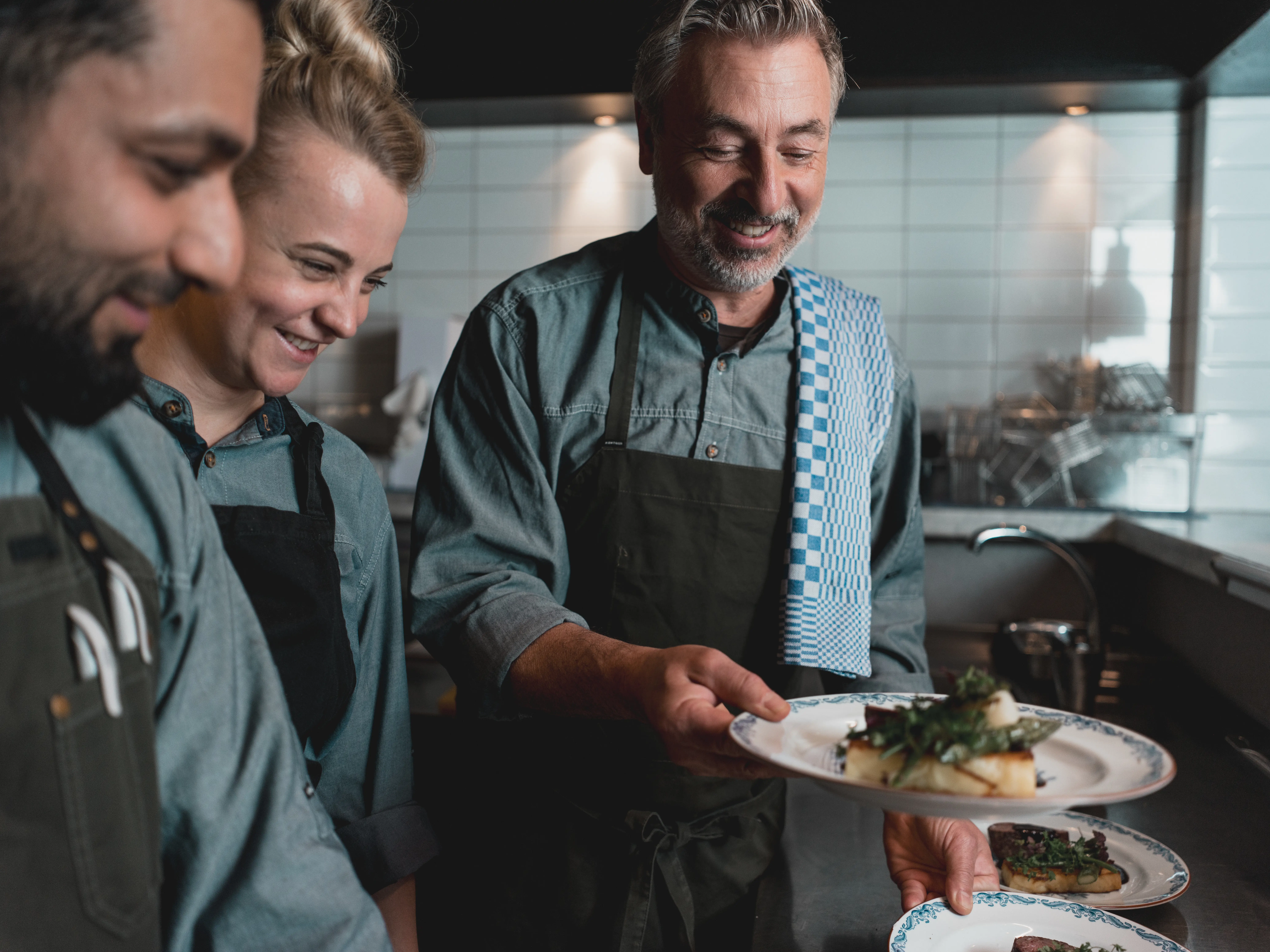 Chef presenting plated dishes to kitchen staff in a modern restaurant kitchen, showcasing teamwork and culinary presentation skills.