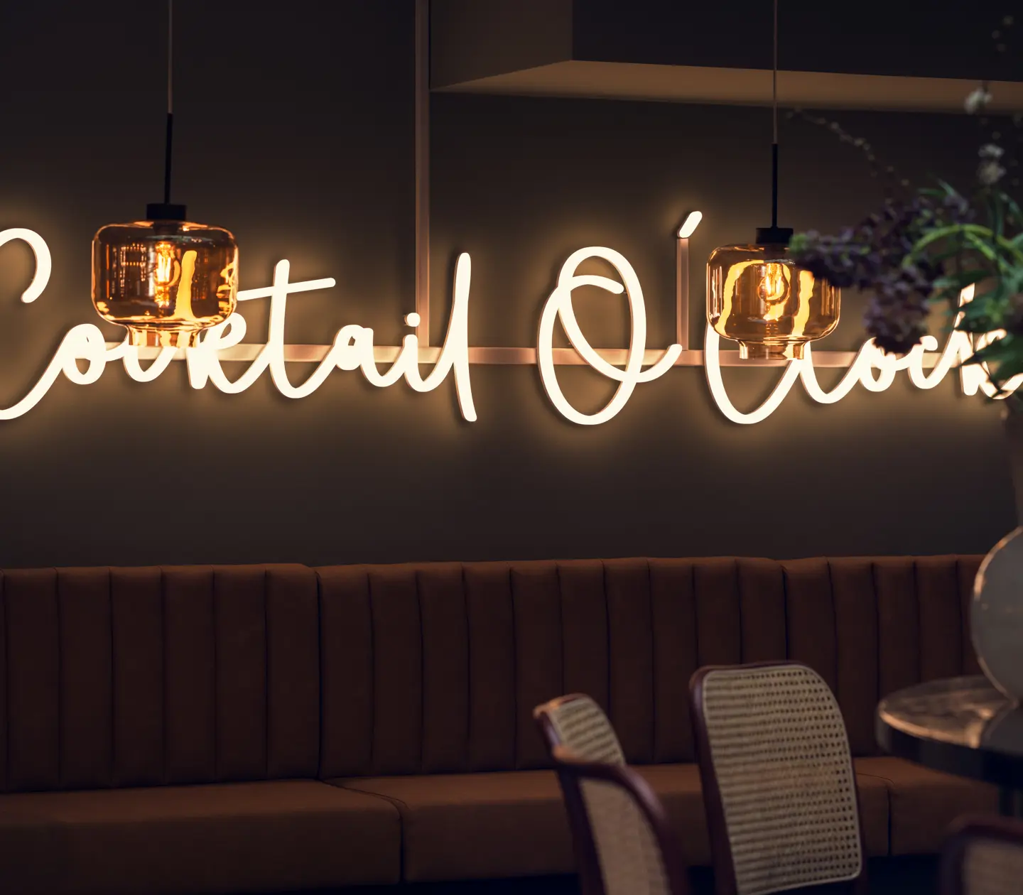 Neon sign reading 'Cocktail Lounge' on a dark wall above a brown sofa in a trendy bar with hanging lights and decorative plants.