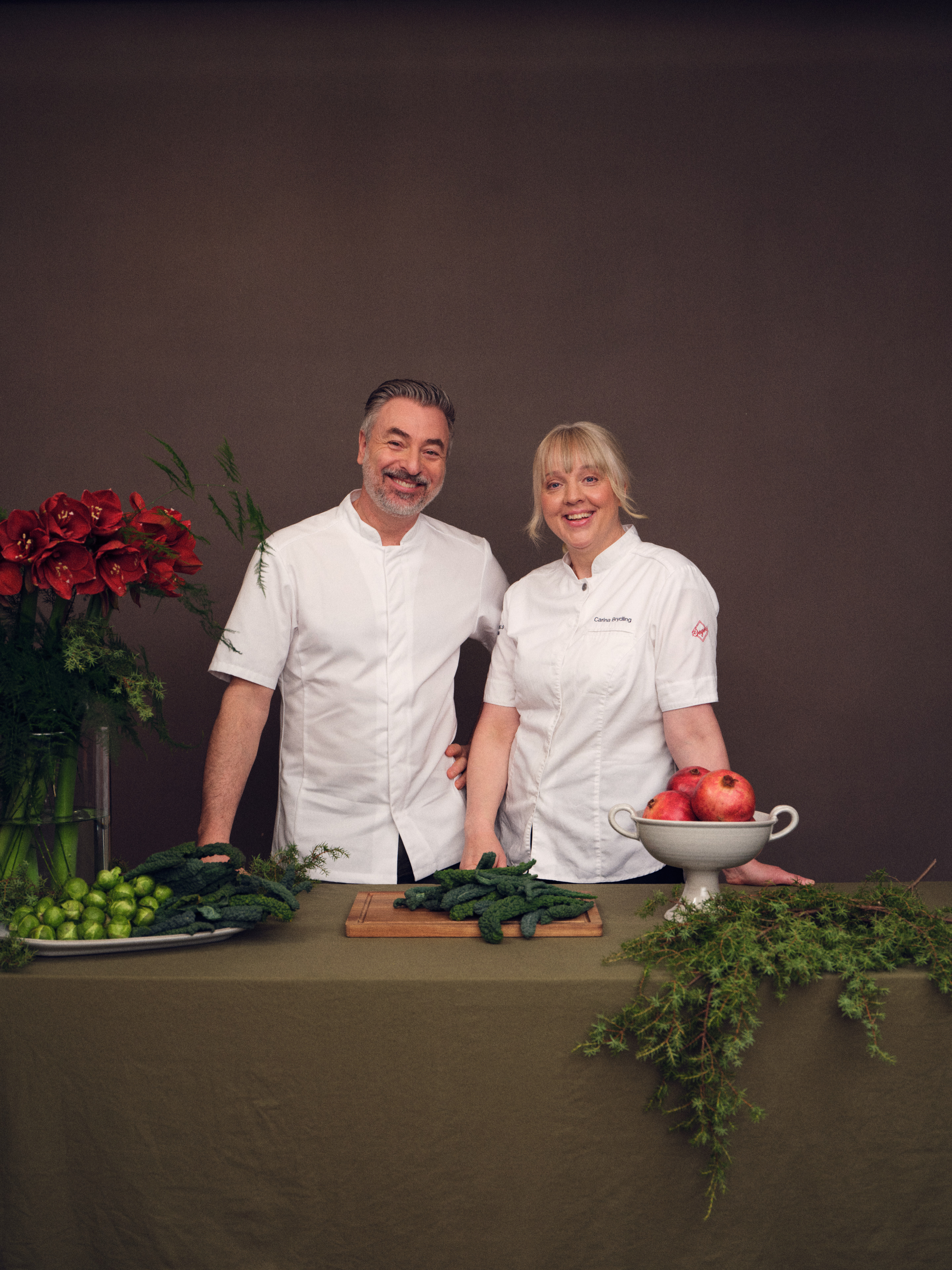 Tareq Taylor & Carina Brydling in white uniforms stand smiling at a festive holiday table adorned with candles, greenery, and a decorated Christmas tree in the background.