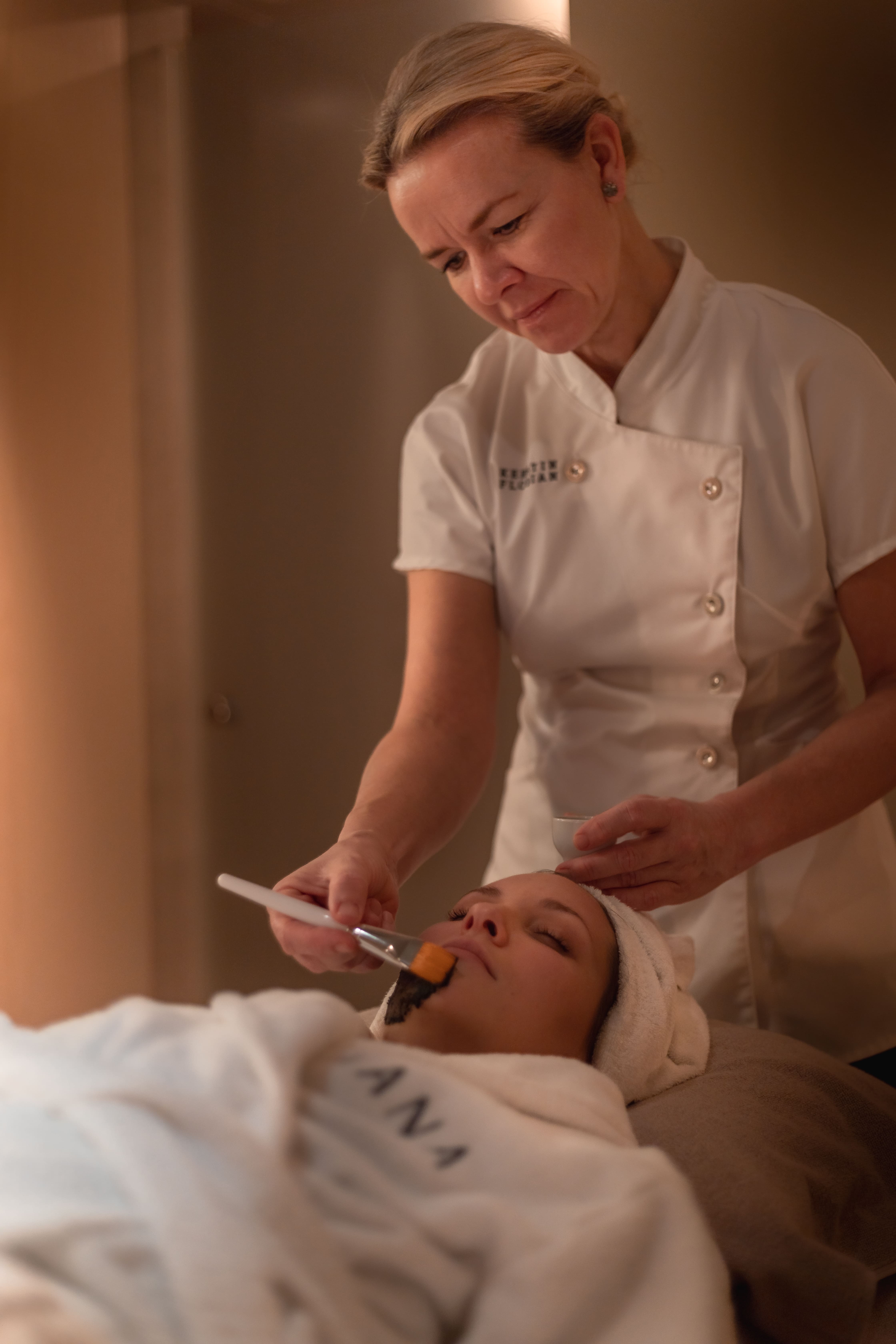 A woman in a white uniform applies a facial mask to a client at the spa