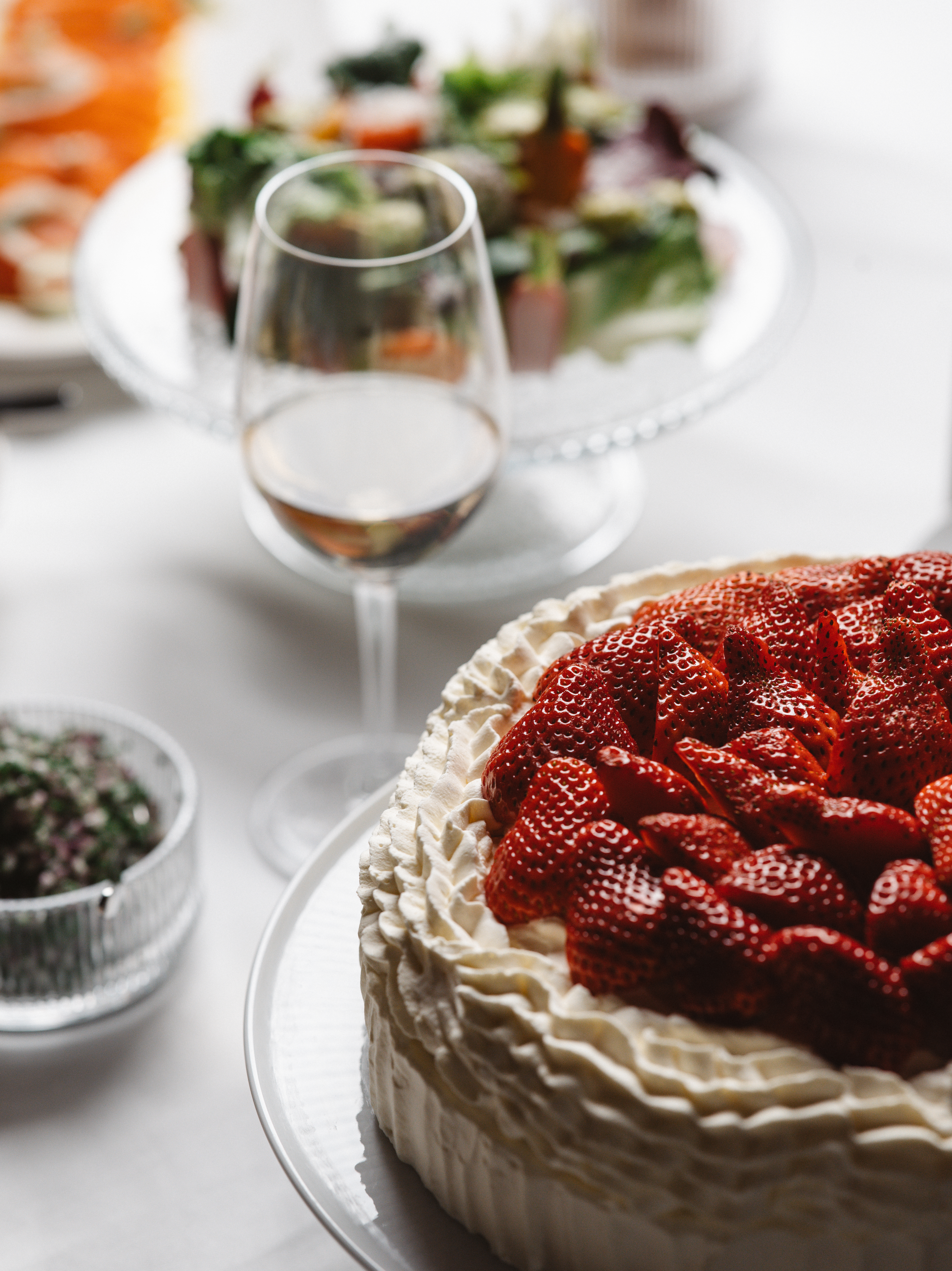 Jordgubbstårta med grädde, serverad på ett bord tillsammans med sallad, vin och andra maträtter.