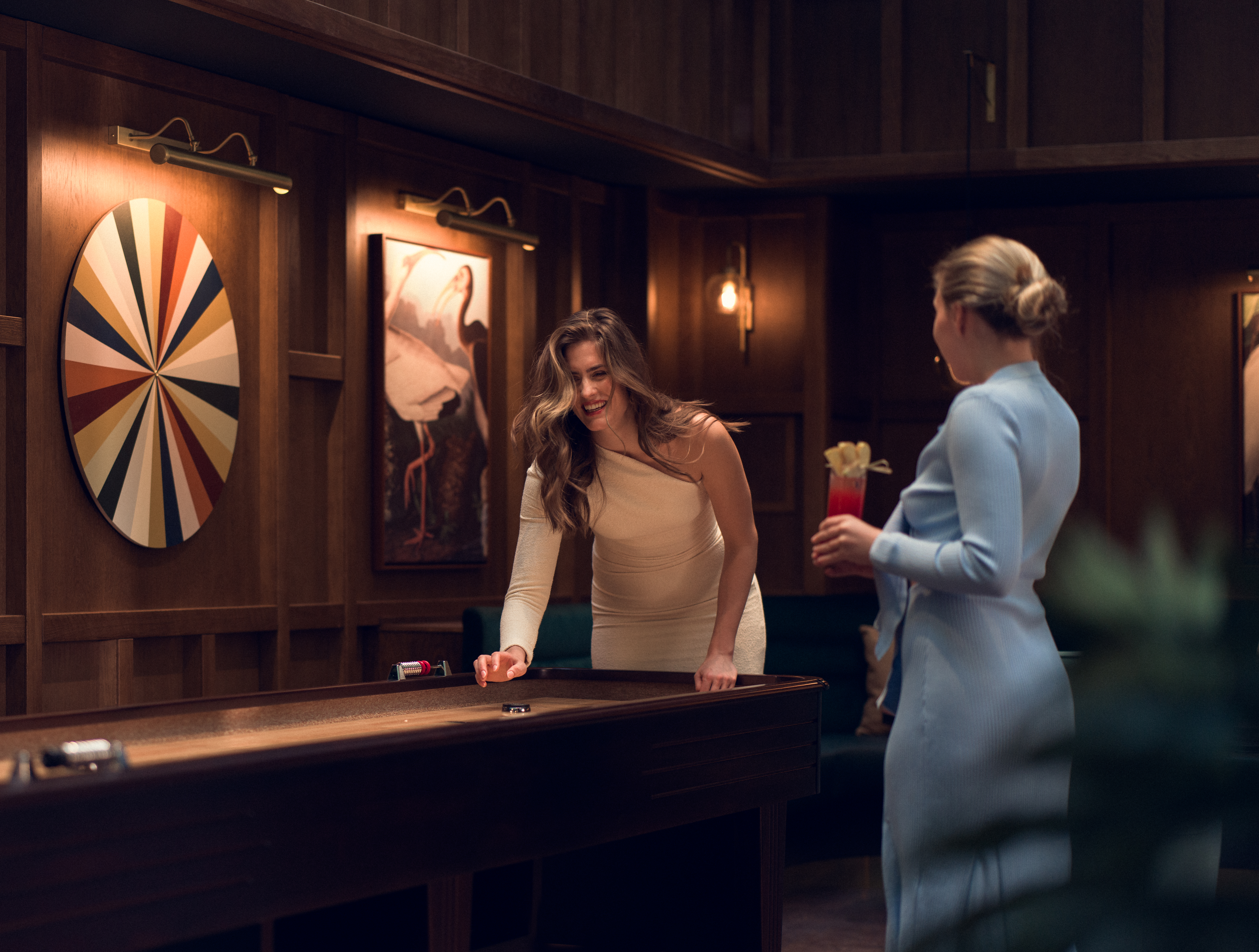 Två kvinnor som spelar shuffleboard i en elegant spelrumsmiljö med darttavla och konst på väggarna.