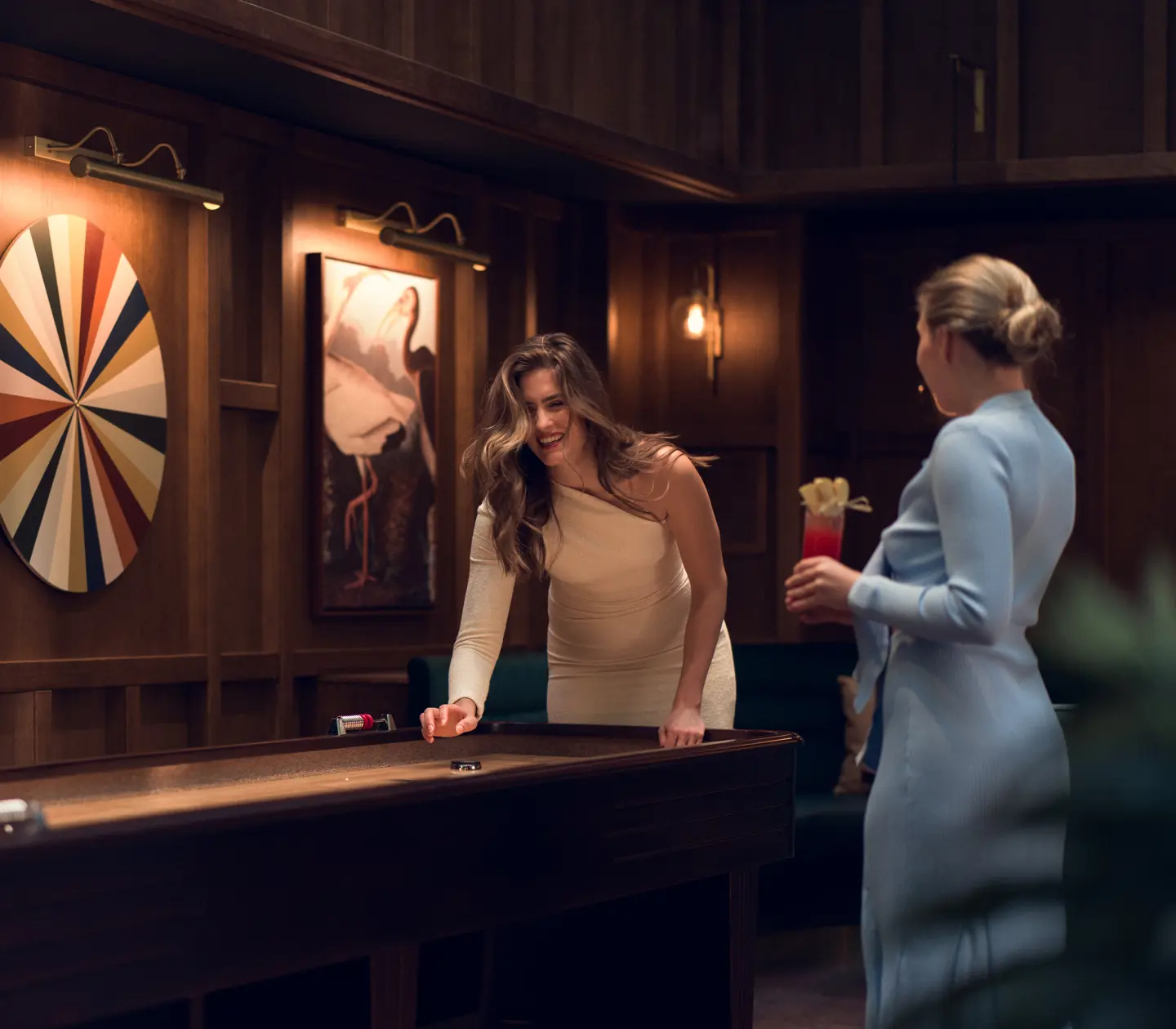 Two women playing shuffleboard in an elegant game room environment, with a dartboard and artwork on the walls.