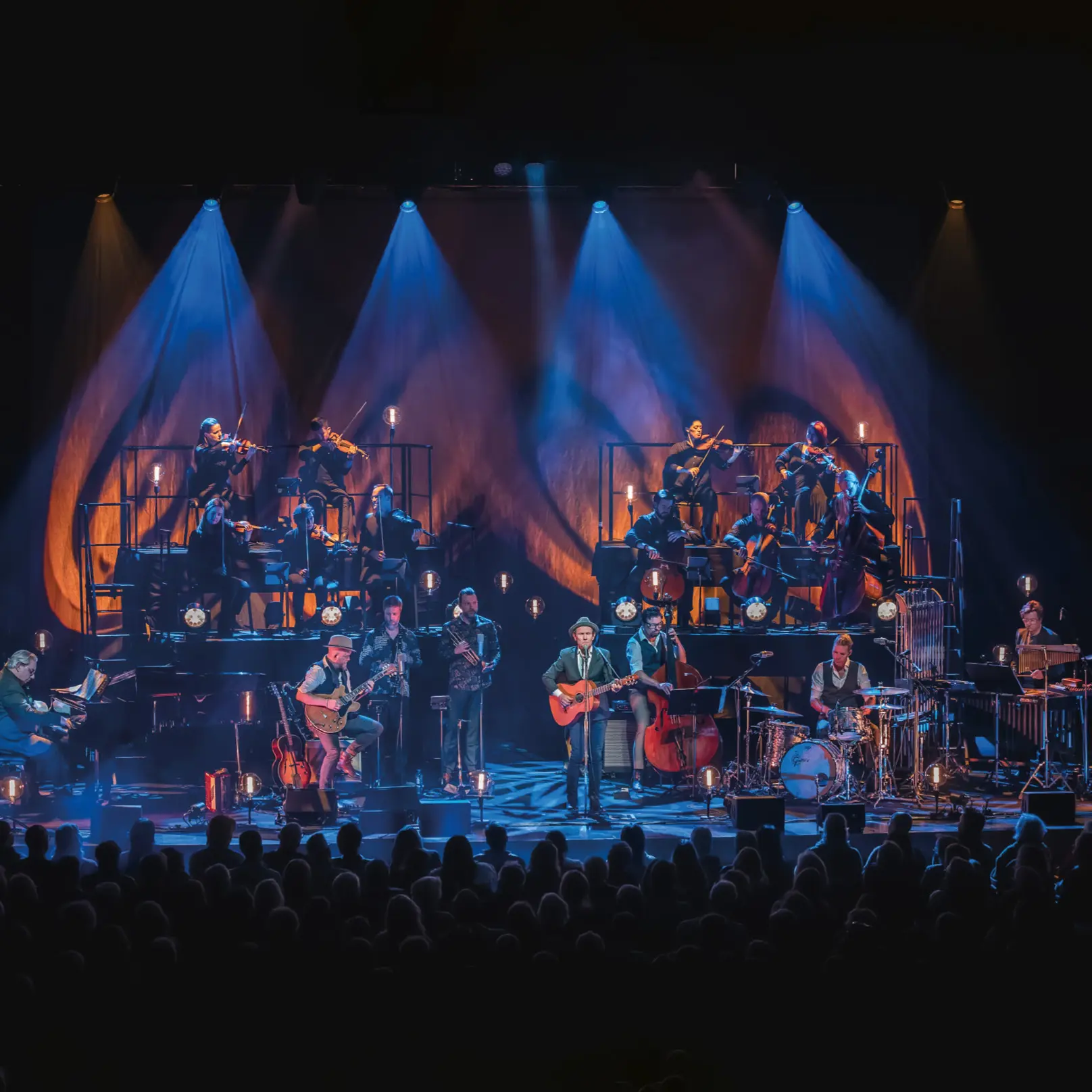 Musikensemble på scen med orkester och band, upplyst av blå scenljus under en livekonsert.