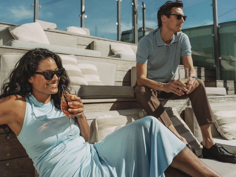 Två personer på utomhusterrass med drycker i solen.