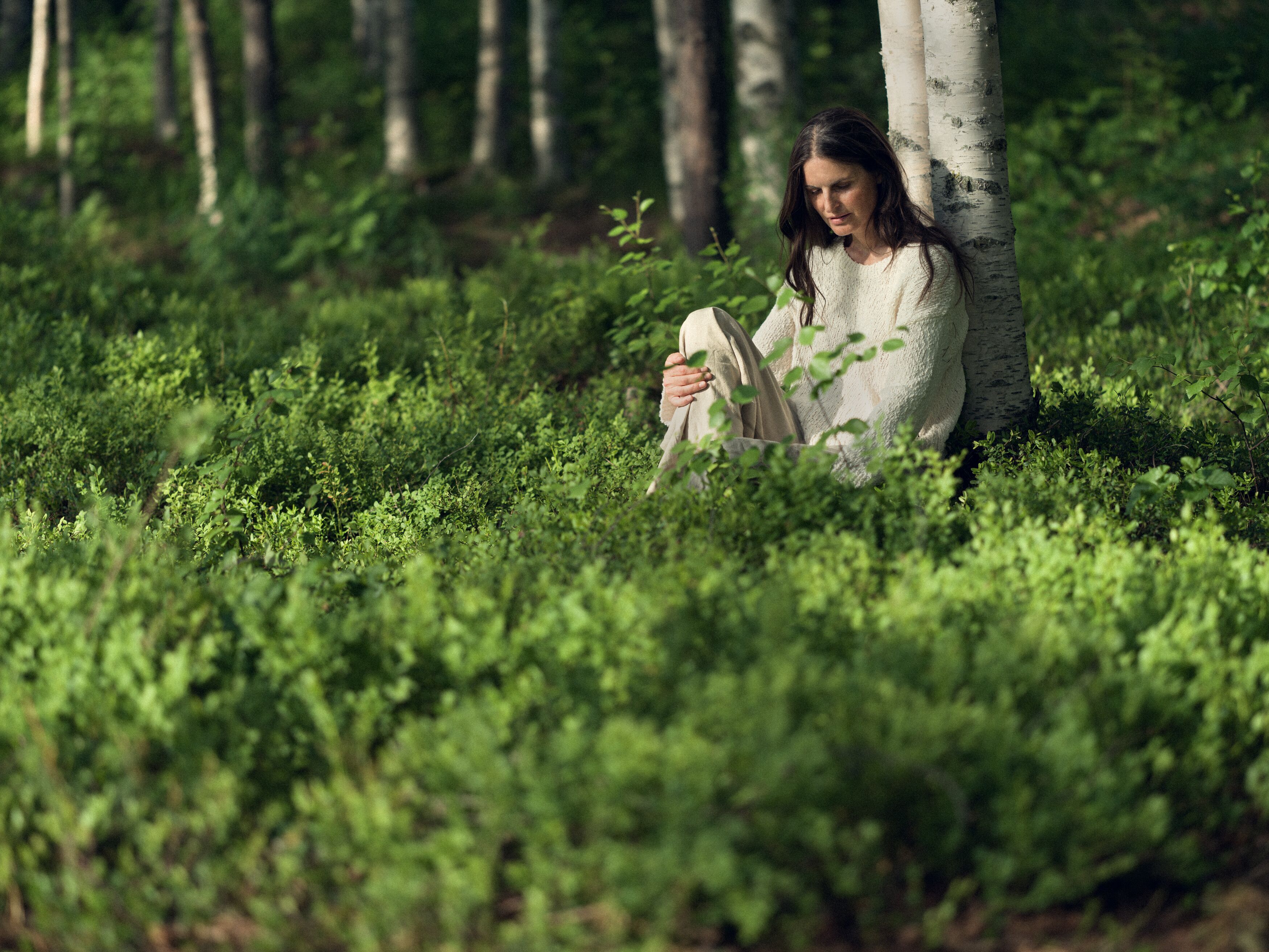 Kvinna i vit tröja sitter lutad mot ett träd i en grön skog.