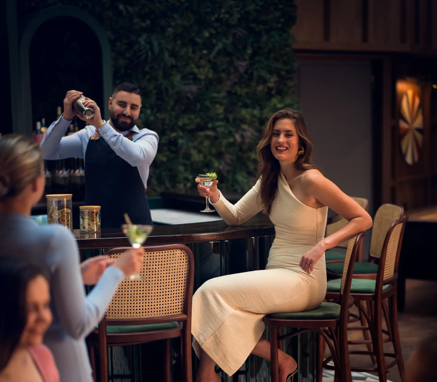 Woman smiling with a drink at an elegant bar while a bartender mixes cocktails in the background.