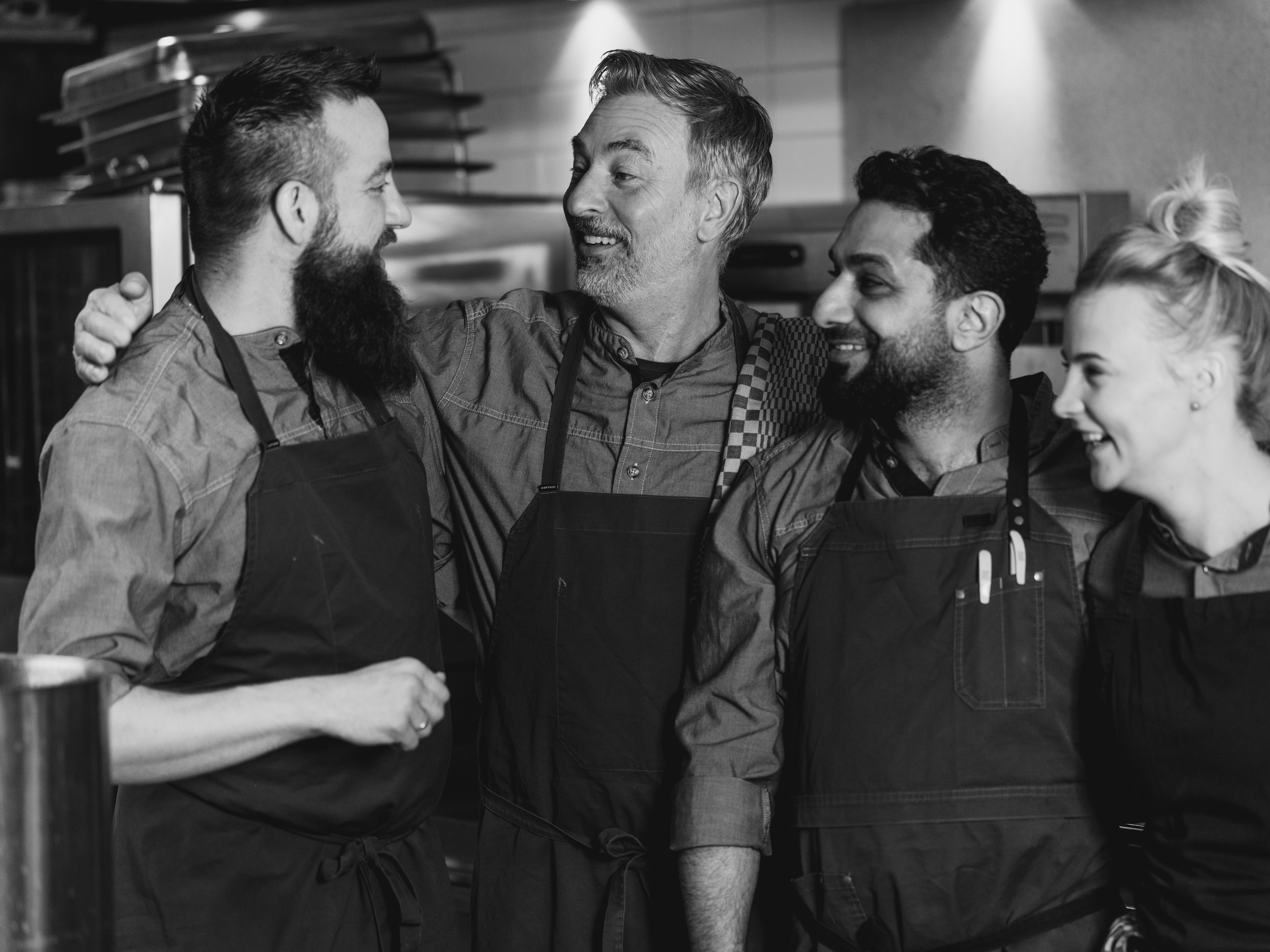 Chefs in a modern kitchen preparing dishes with garnish, wearing gray uniforms and aprons, showcasing teamwork and culinary skills.