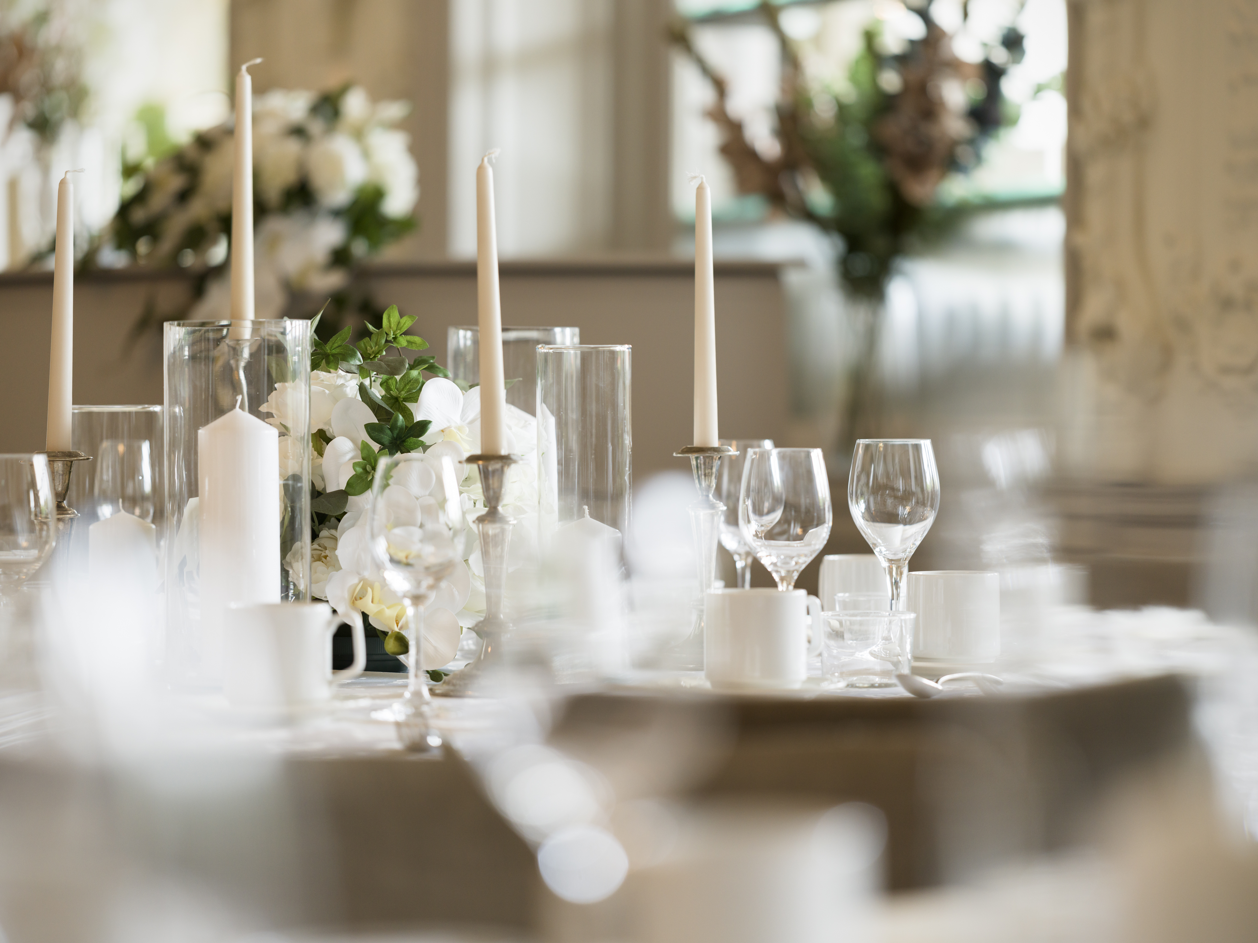 Elegant dukning med vita ljus, porslin och vinglas på ett bröllopsbord, dekorerat med blomsterarrangemang i en ljus och romantisk miljö.