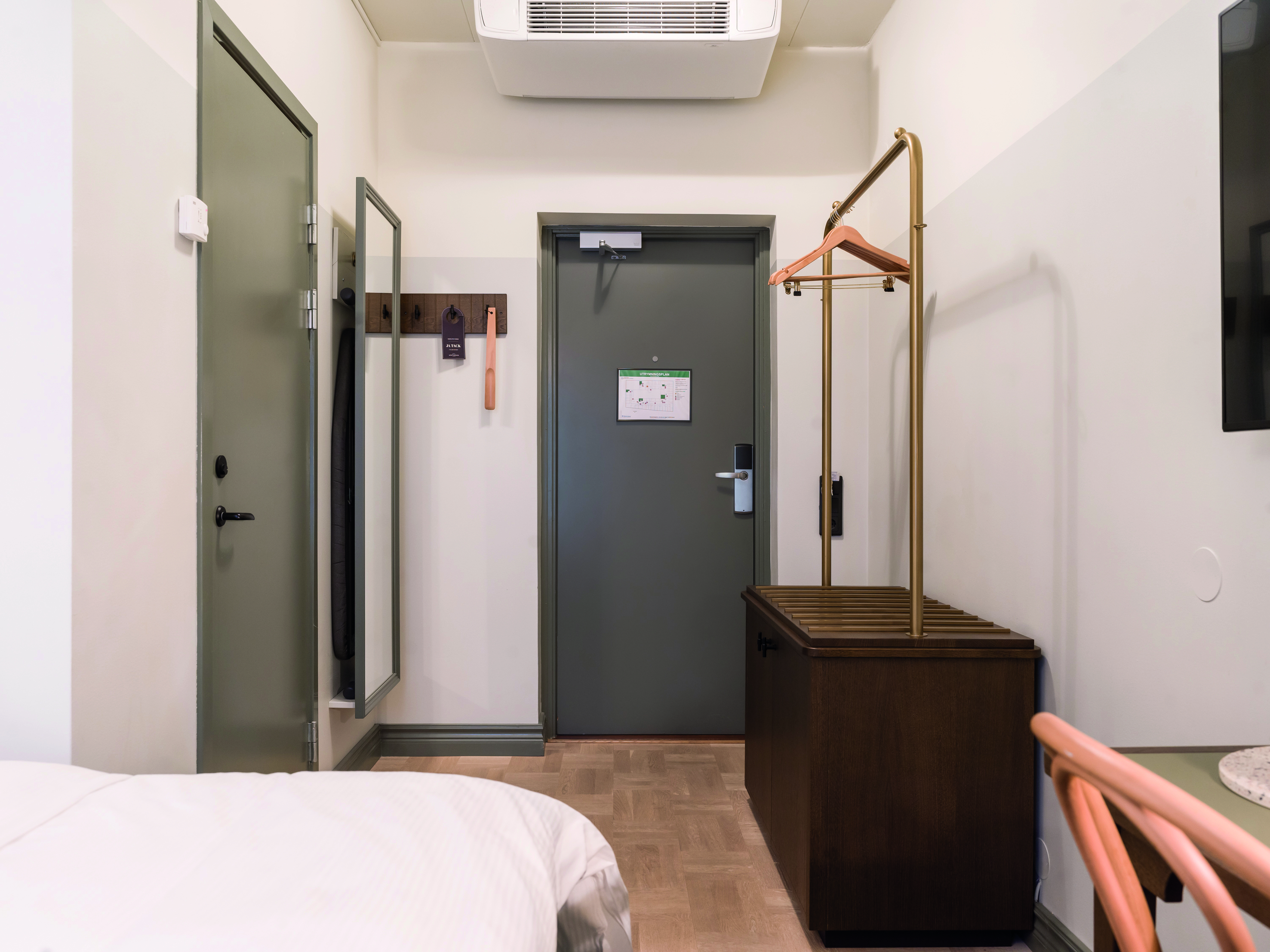 Hotellrumsentré med grå dörr, spegel, klädhängare och träbänk, ljusa väggar och parkettgolv