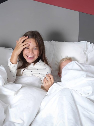 Barn i pyjamas nerbäddade i hotellsäng med vita lakan