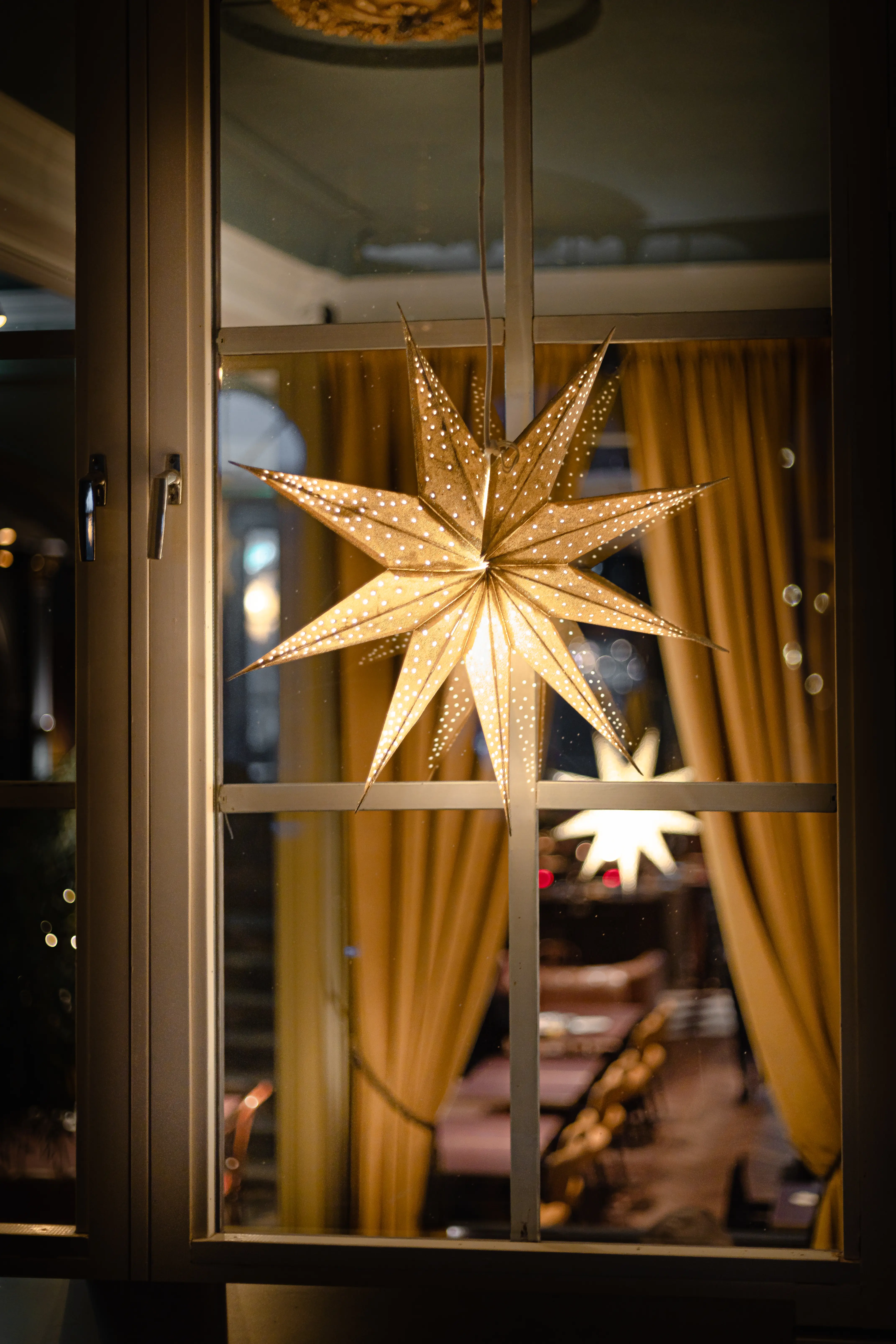 Illuminated paper star lantern hanging in a window with warm yellow curtains, creating a cozy indoor ambiance.