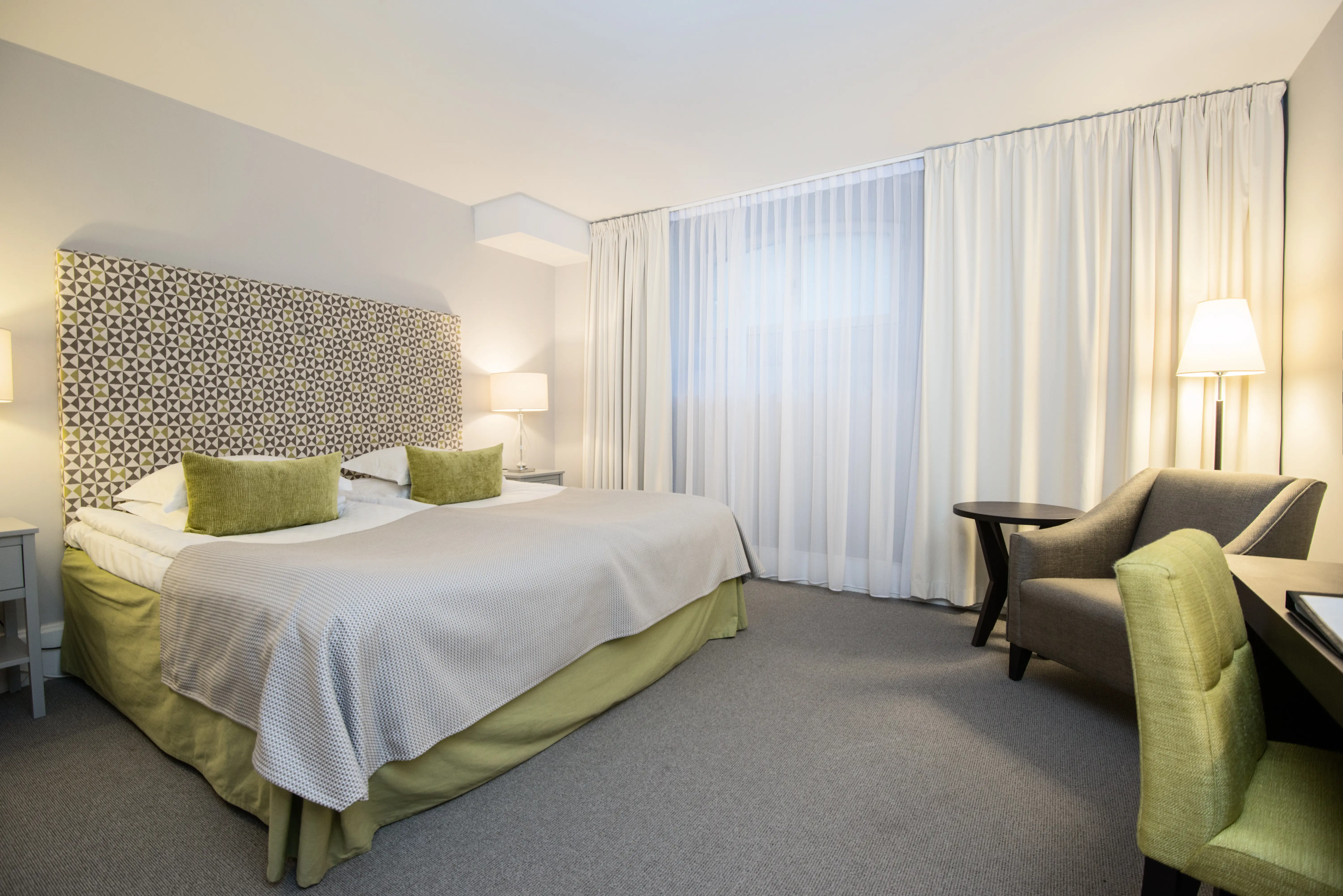 Ljust hotellrum med säng, fåtölj och gardier