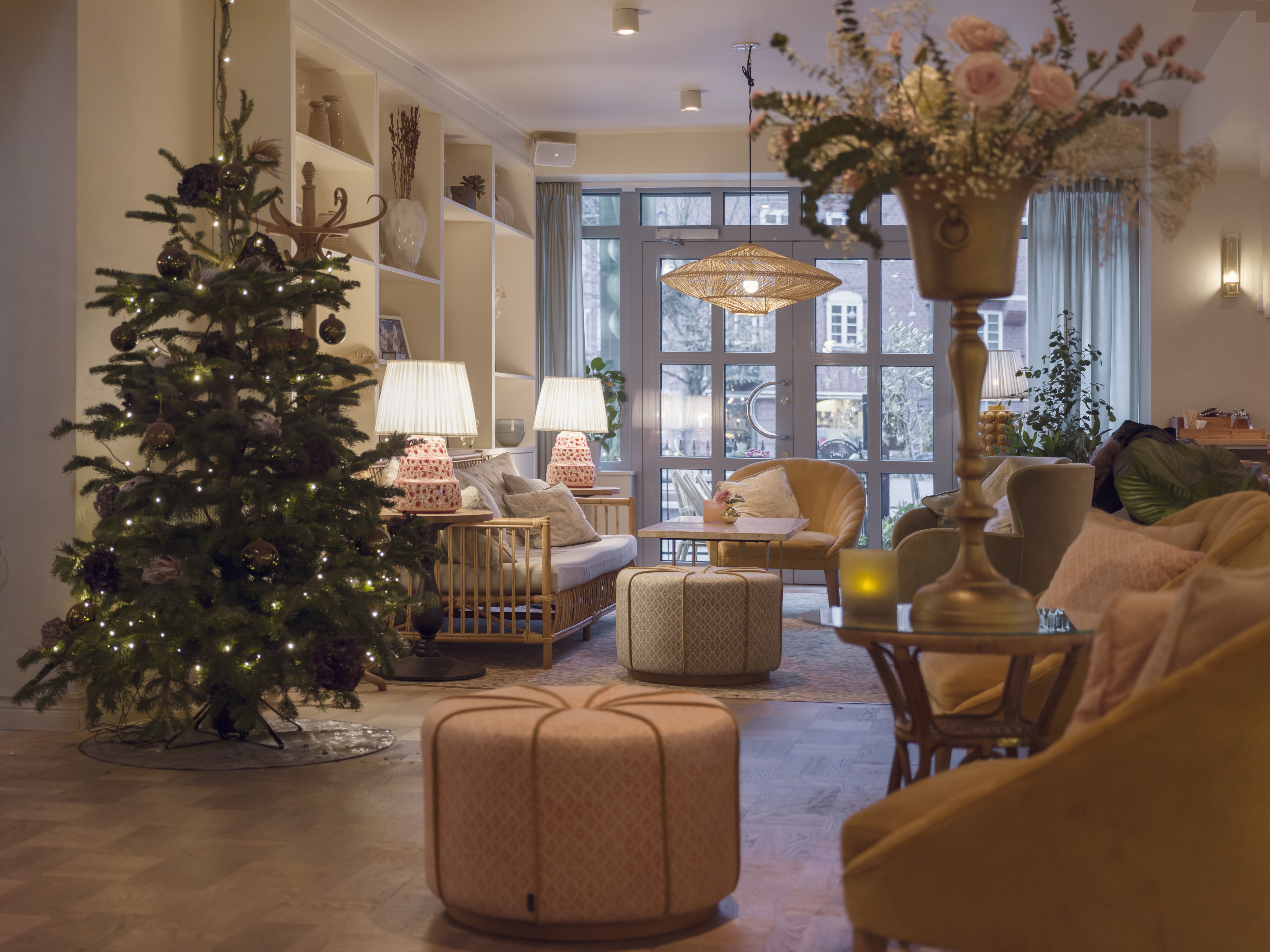 Juldekorerat vardagsrum med en gran klädd i ljus, moderna soffor och fåtöljer, harmoniskt belysta bordslampor och blommor på ett bord i förgrunden.