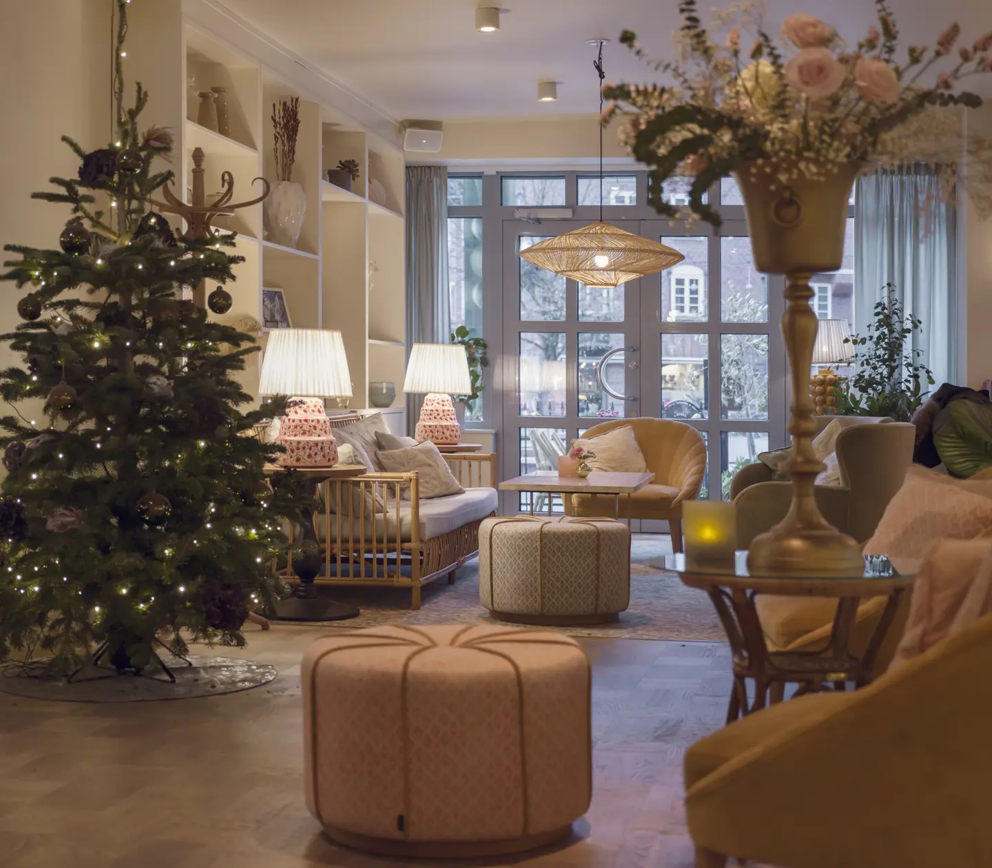 Juldekorerat vardagsrum med en gran klädd i ljus, moderna soffor och fåtöljer, harmoniskt belysta bordslampor och blommor på ett bord i förgrunden.