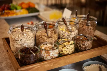 Glasburkar med olika typer av müsli, flingor, torkad frukt och nötter på en träbricka, del av en frukostbuffé