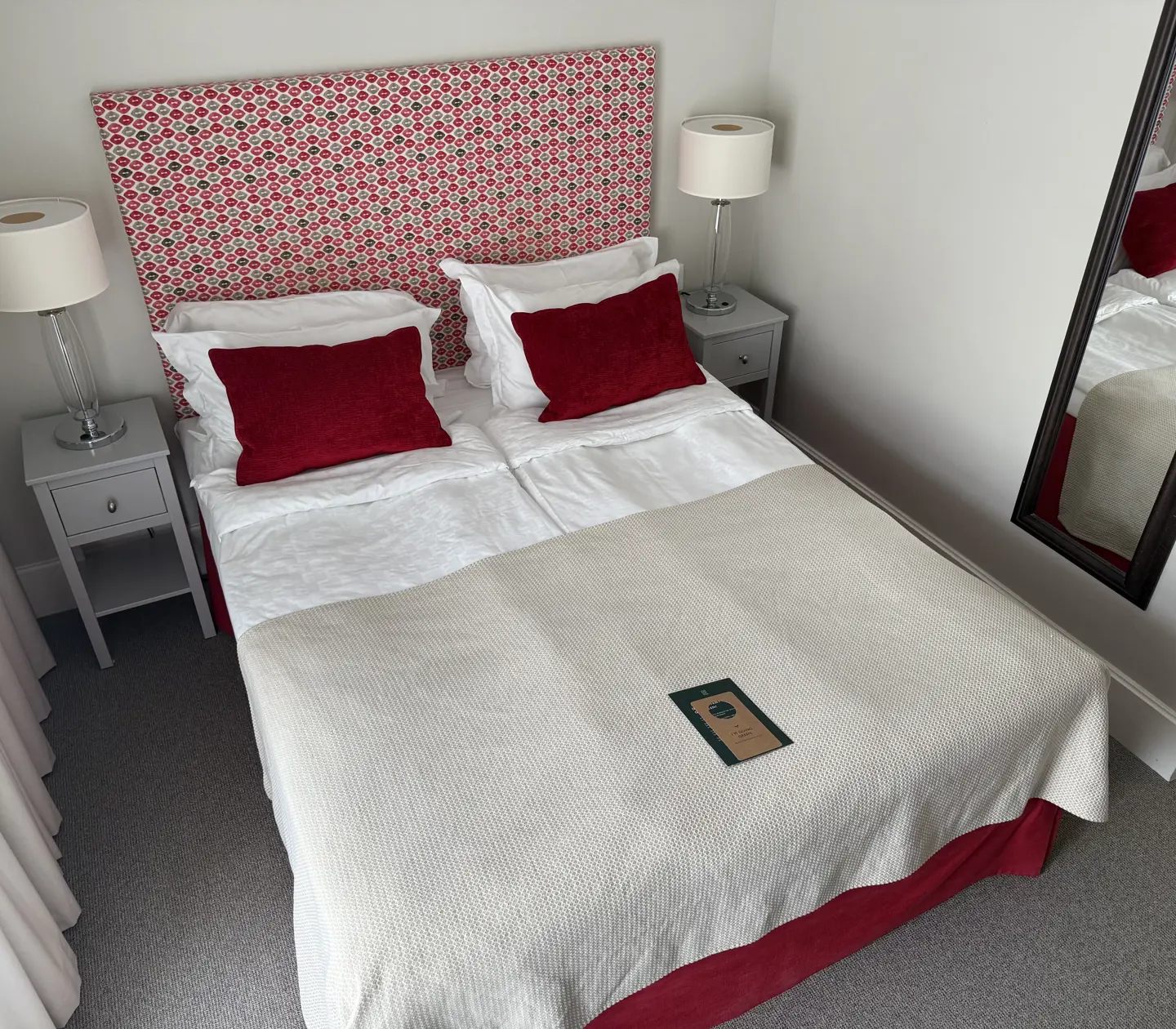 Hotellrum med bäddad säng, rött och vitt överkast samt dekorativa kuddar. Sänglampor står på vita nattduksbord.