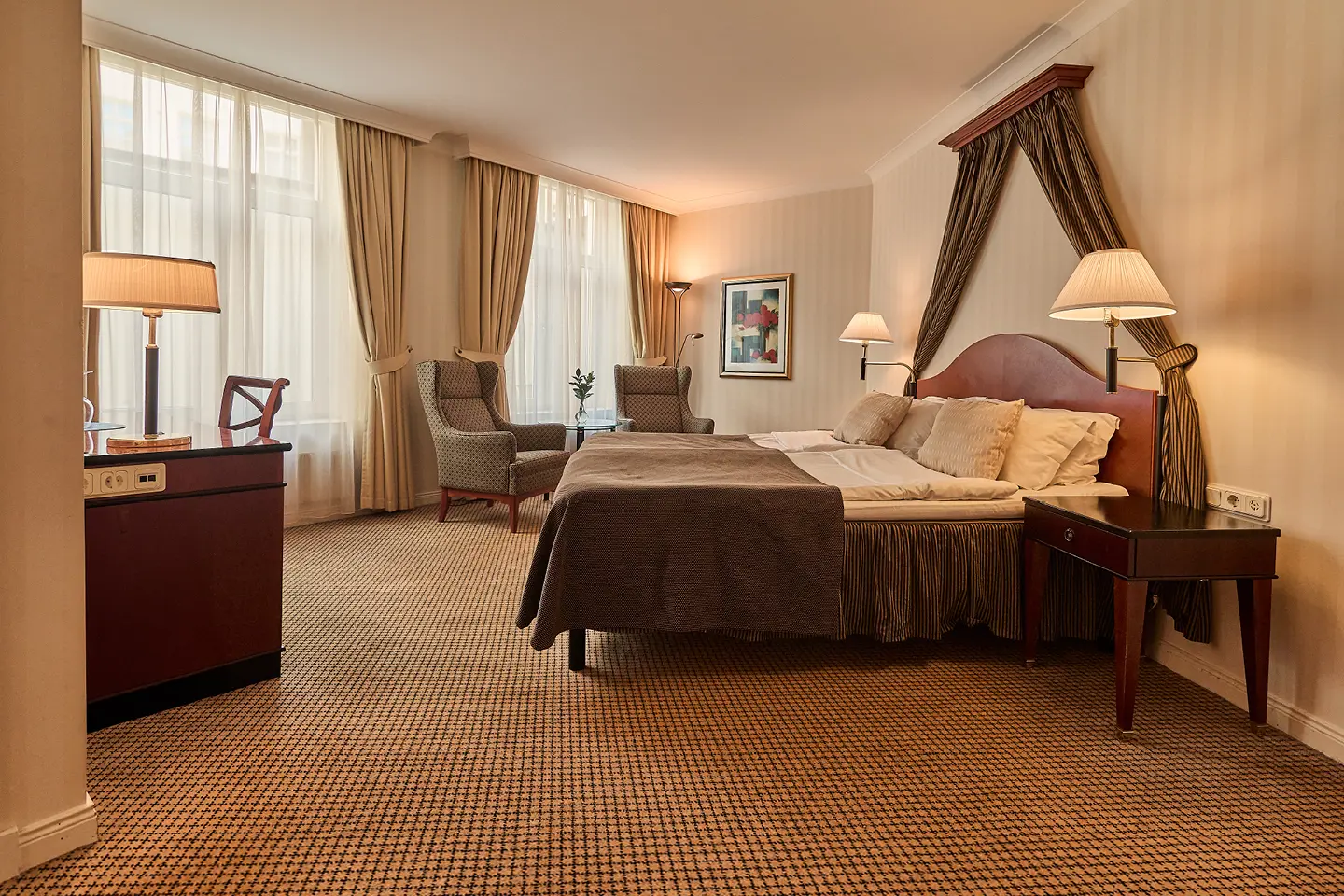 Modern hotel room with a cozy bed, stylish gray armchair, comfortable sofa, and round glass coffee table, accented by framed art and large windows with beige curtains.