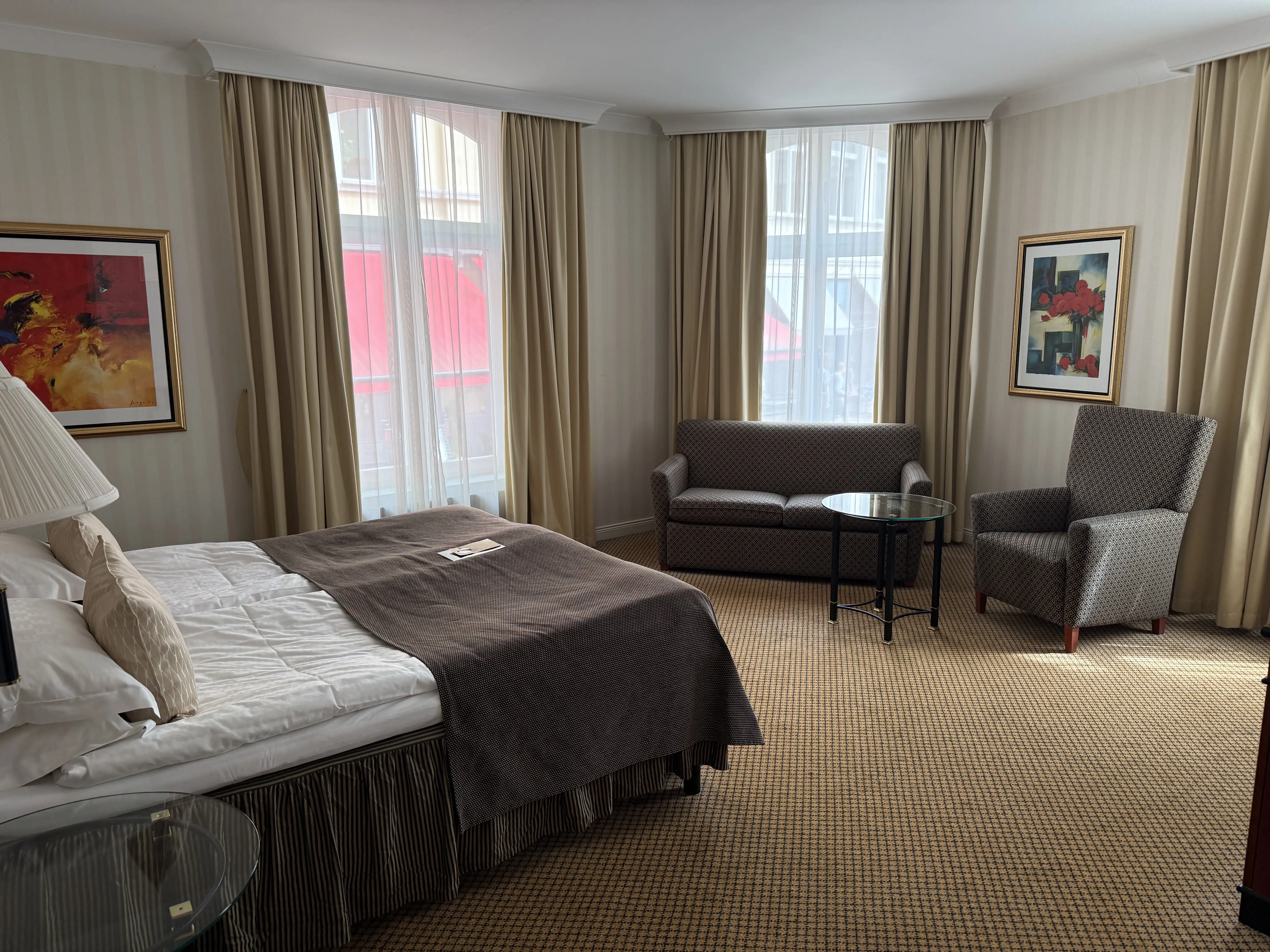 Hotellrum med en stor säng, soffa, fåtölj och konstverk på väggarna. Naturligt ljus från stora fönster bakom ljusa gardiner.