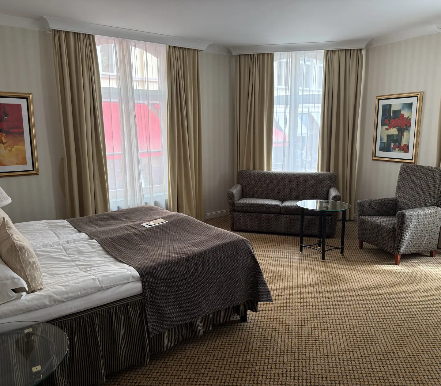 Hotellrum med en stor säng, soffa, fåtölj och konstverk på väggarna. Naturligt ljus från stora fönster bakom ljusa gardiner.