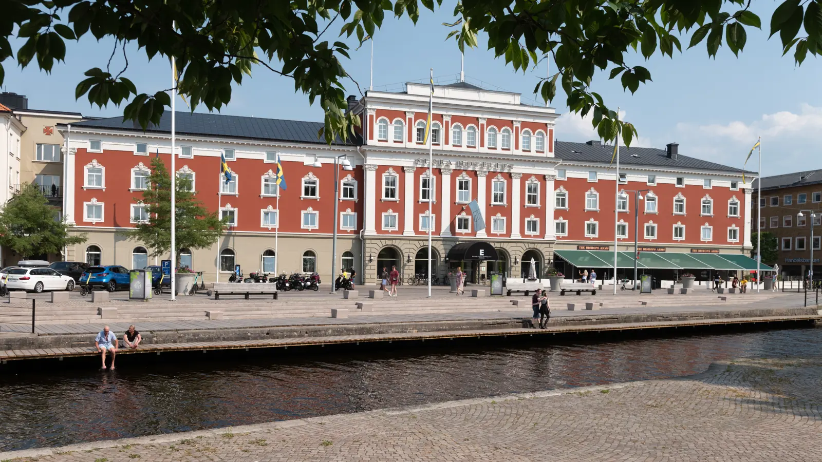Rött historiskt hotell vid en kanal i centrala Jönköping, Sverige, med folk som promenerar och sitter vid vattnet under en klar himmel