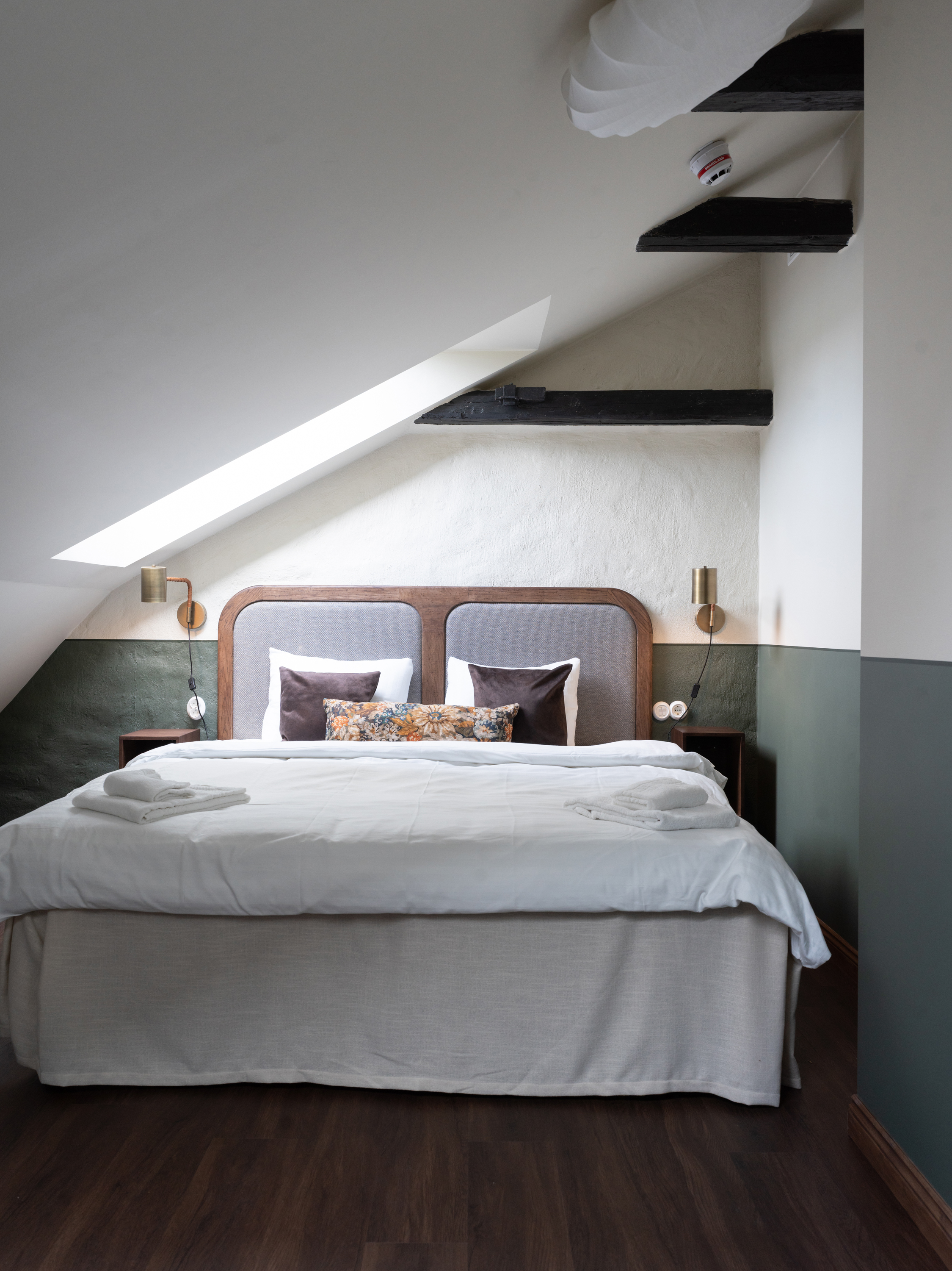 Snyggt sovrum med snedtak, modern säng med vita och bruna kuddar, vita lakan och två handdukar, trälaminatgolv, vägglampor och mörka hyllor i hörnet.