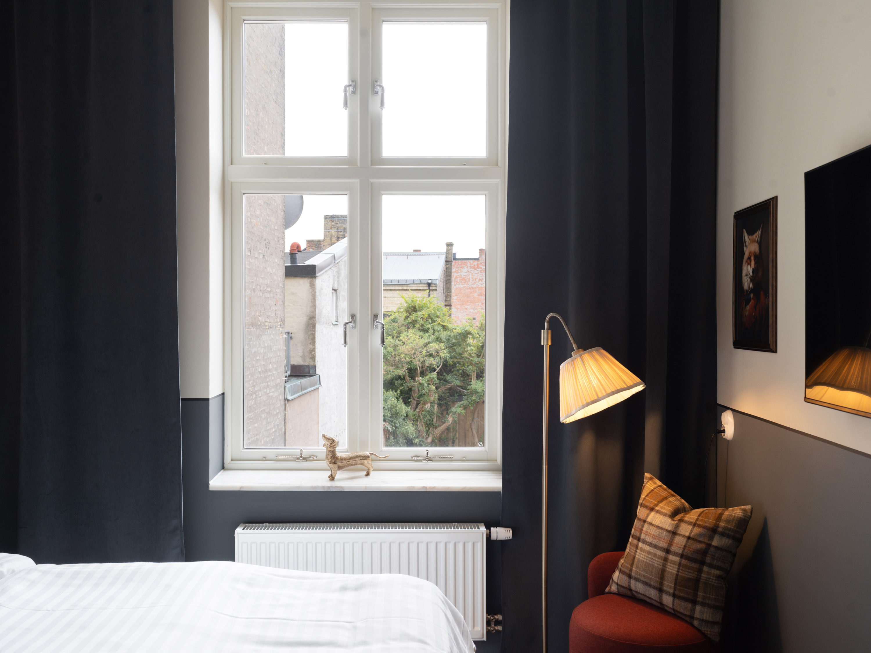 Sovrum med stort fönster och utsikt mot innergård, mörka gardiner, säng med vit bäddning, golvlampa och väggdekor med djurmotiv.