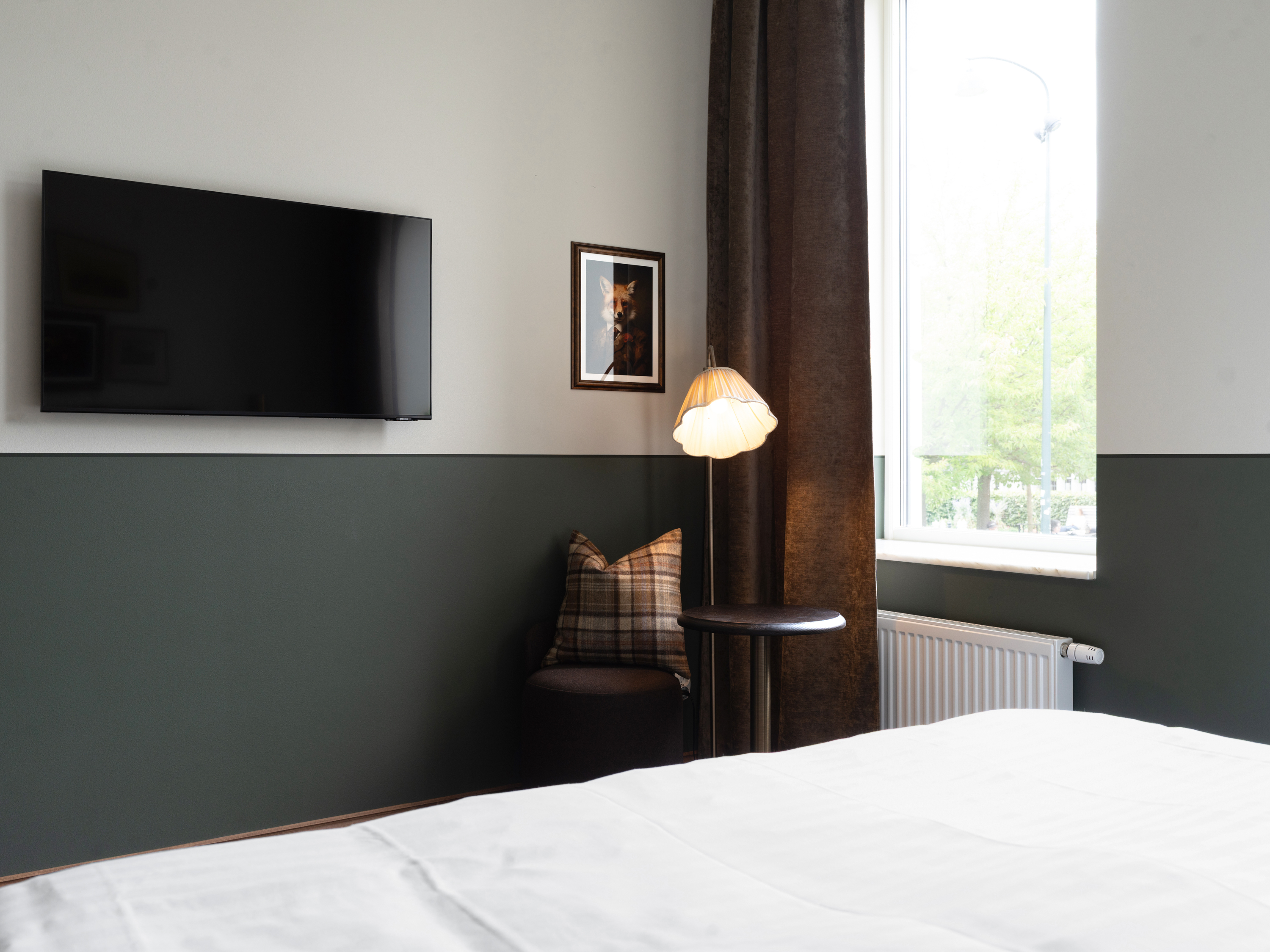 Ljust och modernt sovrum med en dubbelsäng, två sänglampor, och en träbyrå. Väggarna är gula och vita med en inramad bild på väggen. Fönster med vita gardiner släpper in naturligt ljus, och ett skrivbord med stol står i hörnet.