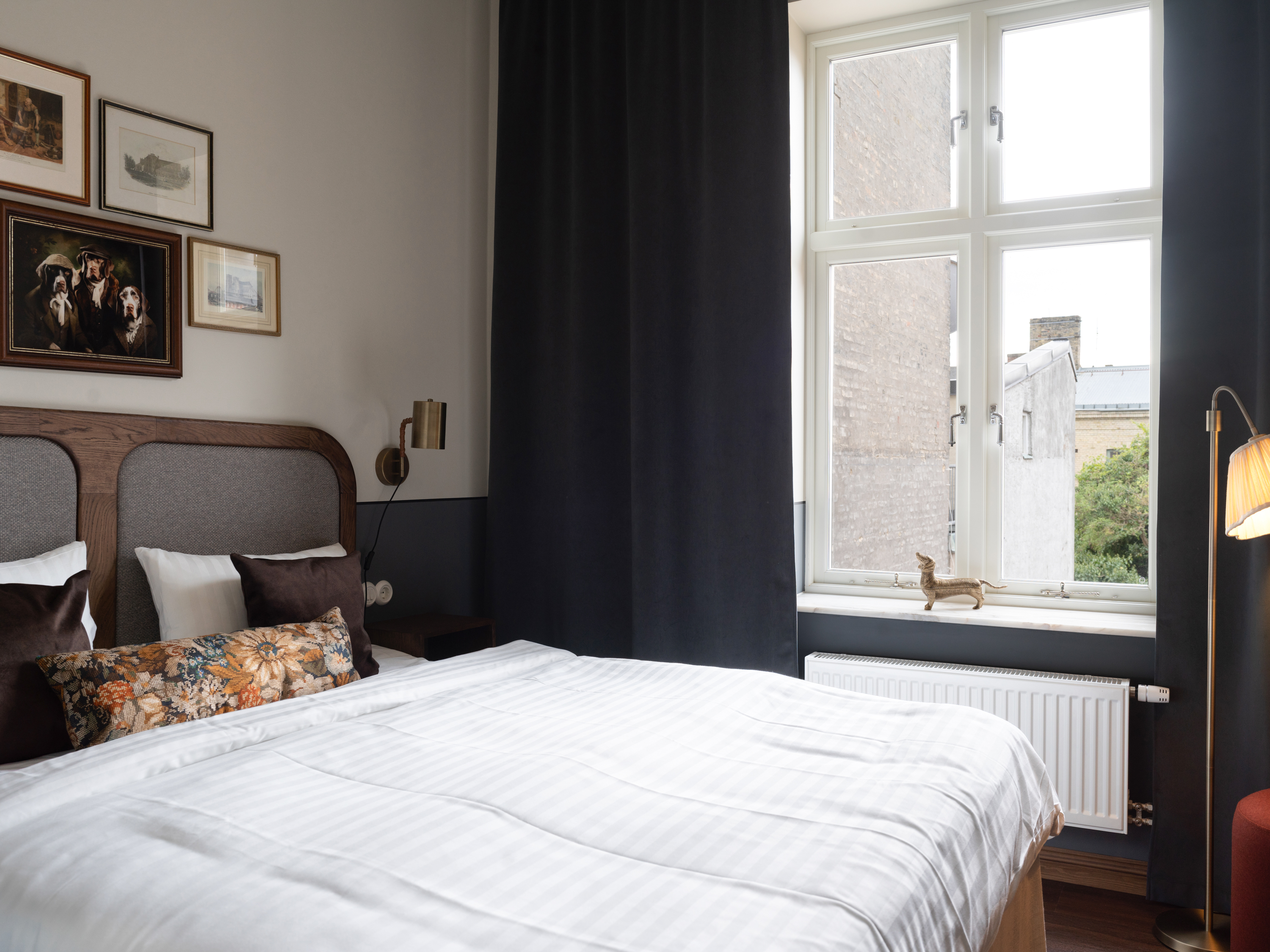 Sovrum med en dubbelsäng, vita sängkläder och prydnadskuddar. Väggkonst ovanför sängen. Ett fönster med mörka gardiner släpper in dagsljus från utsidan.