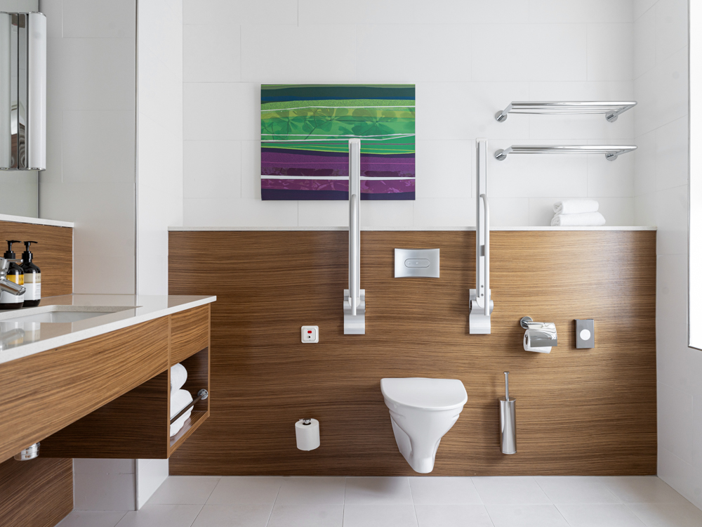 Handikappanpassat badrum med vägghängd toalett, träpanelsdetaljer och stödhandtag, samt modern konst på väggen.