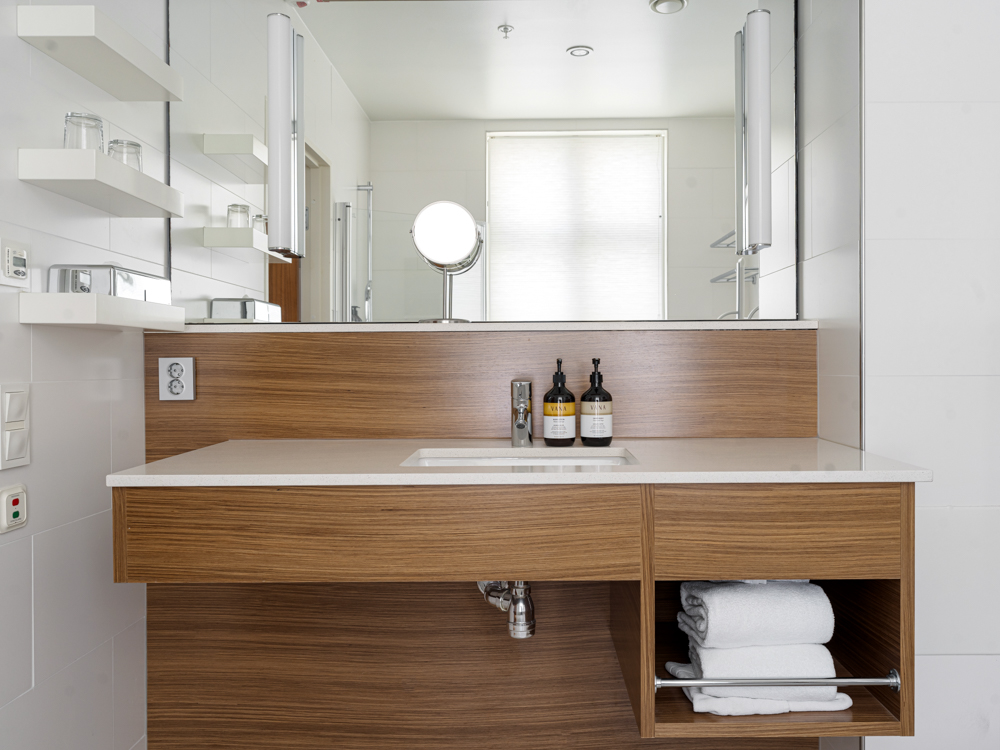 Modernt badrum med träpanel, vit bänkskiva och integrerad spegel. Flaskor står på handfatet och handdukar är prydligt vikta under. Hyllor med glas ovanför eluttag.