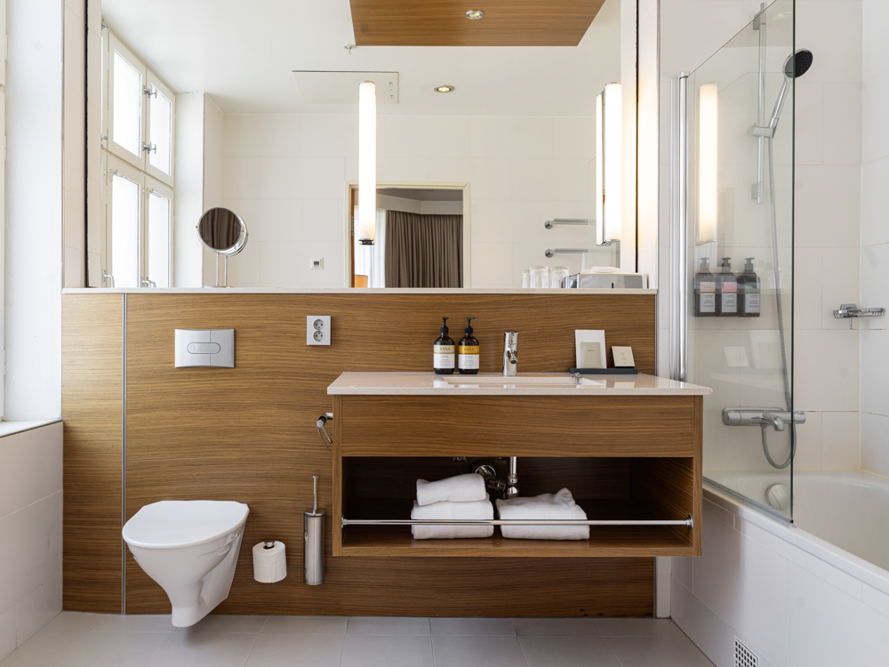 Modernt badrum med träpanel, vägghängd toalett, spegel och öppen dusch med glasvägg.