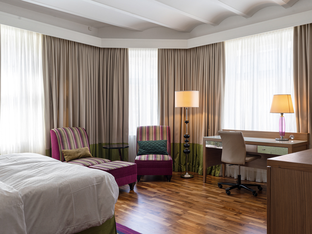 Hotellrum med modernt skrivbord, fåtölj och golvlampor, träparkett och stora fönster med ljusa gardiner.