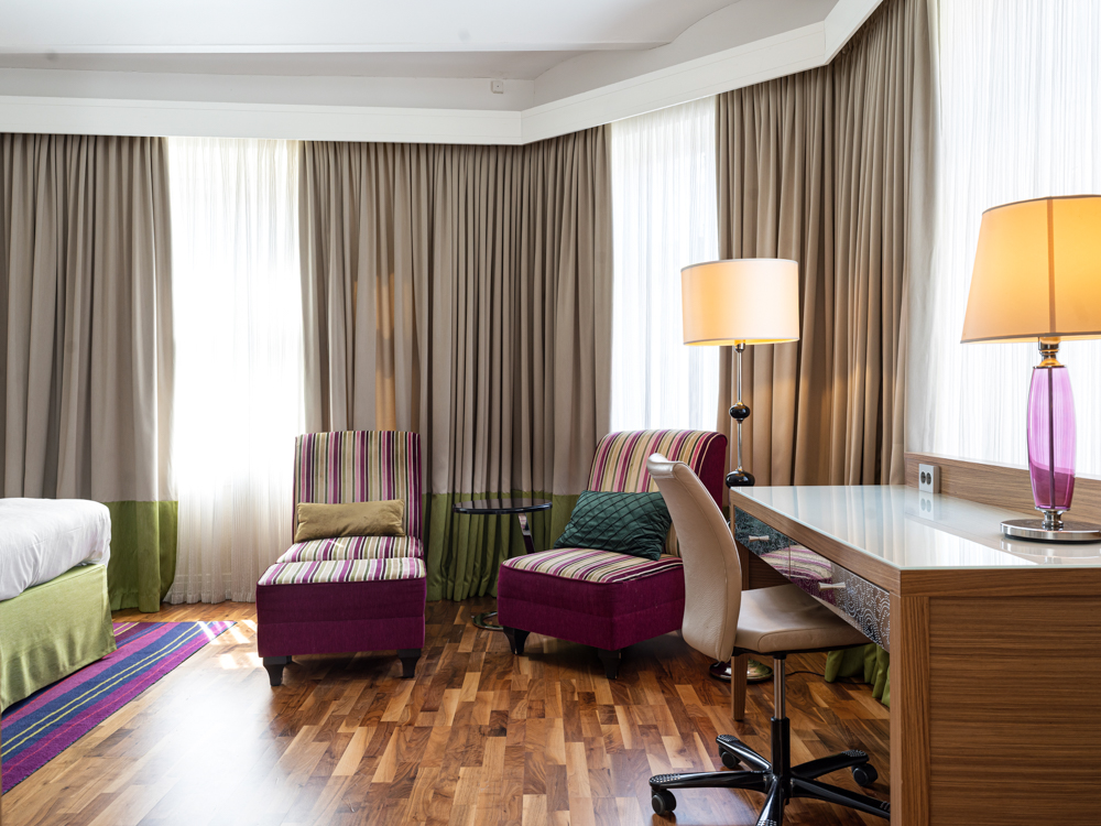 Modernt hotellrum med träparkett, två färgglada fåtöljer, skrivbord och lampa.