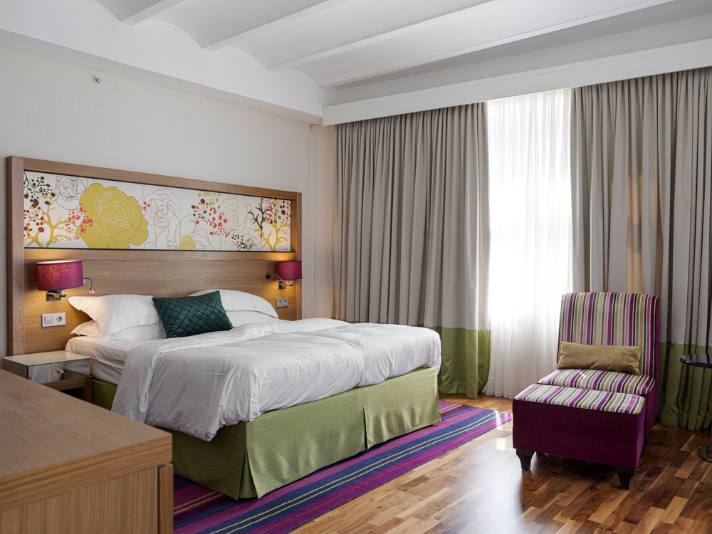 Modernt hotellrum med en dubbelsäng, blommönstrad huvudgavel och dekorativa lampor. Rummet har trägolv, gardiner och en färgglad fåtölj med randigt mönster.