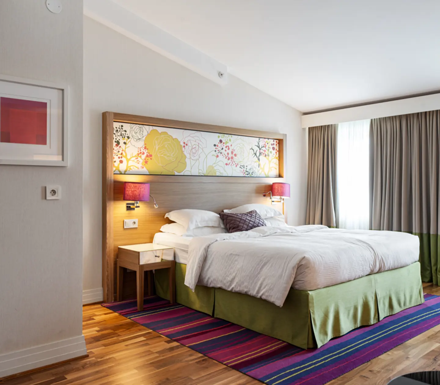 Hotellrum med dubbelsäng, färgglad huvudgavel och gardiner, ljus och modern inredning.