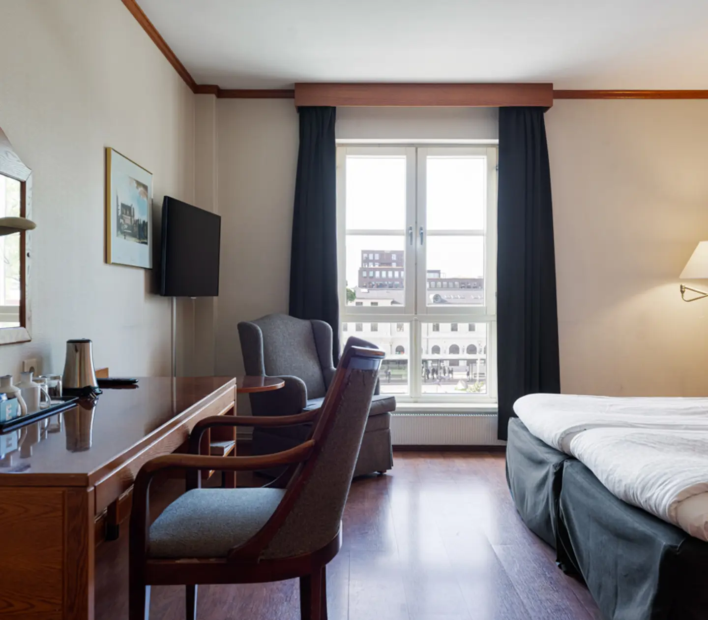 Cozy hotel room with a large bed, wooden desk, and armchair, featuring a window view of city architecture.