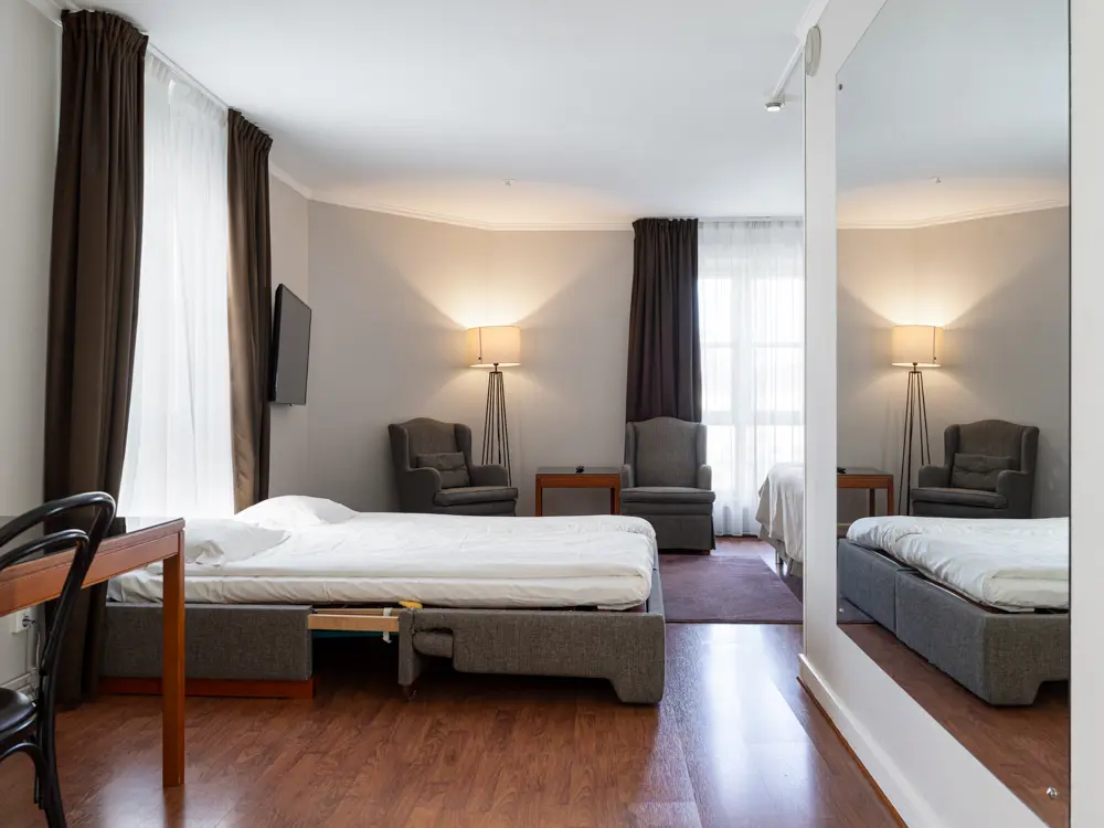 Contemporary hotel room with a single bed, cozy armchair, floor lamp, and large mirror reflecting the space. Room features neutral colors and wood flooring with natural light from large window.
