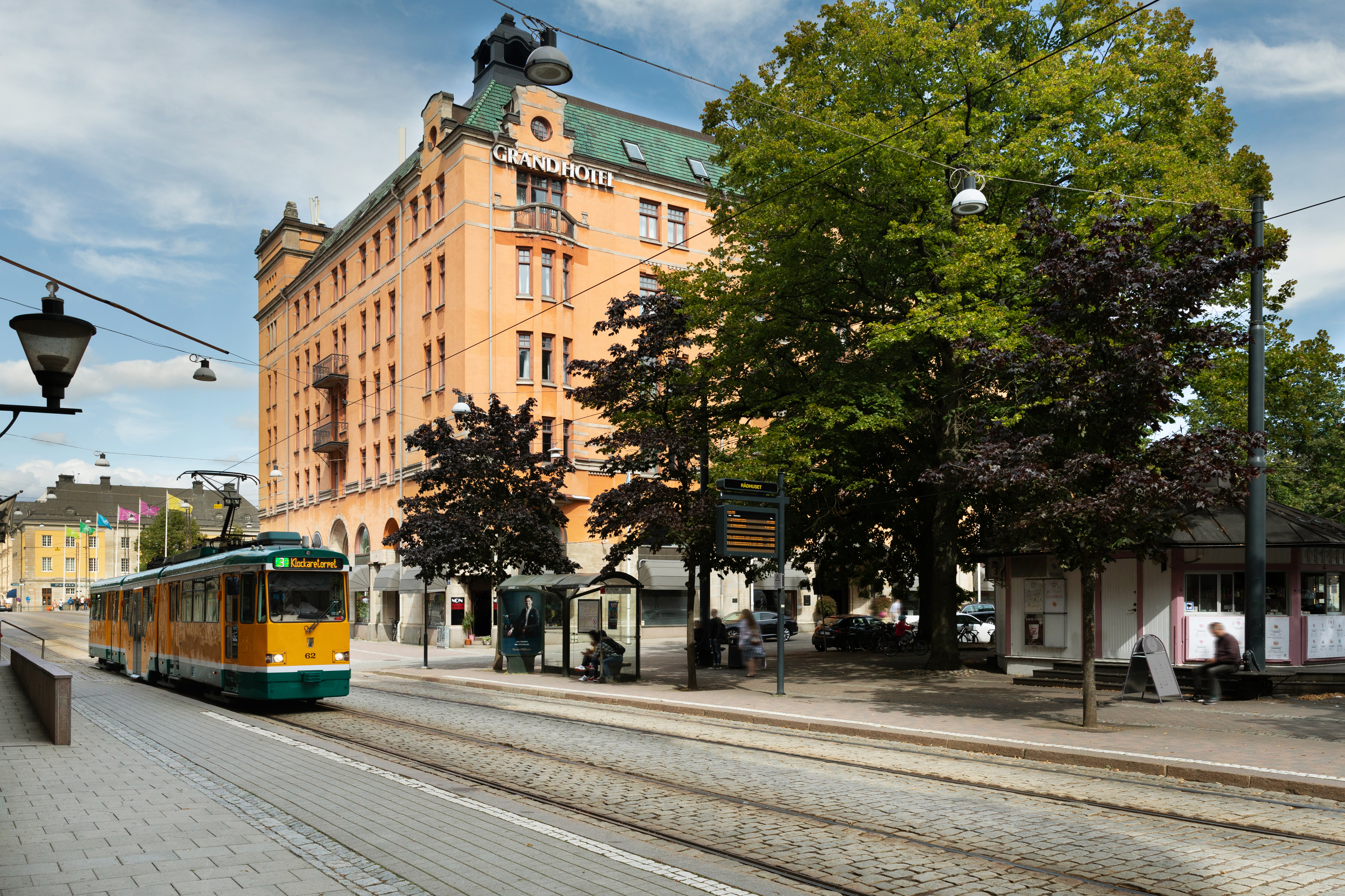 The orange facade of the Elite Grand Hotel in Norrköping with trees and a tram in front